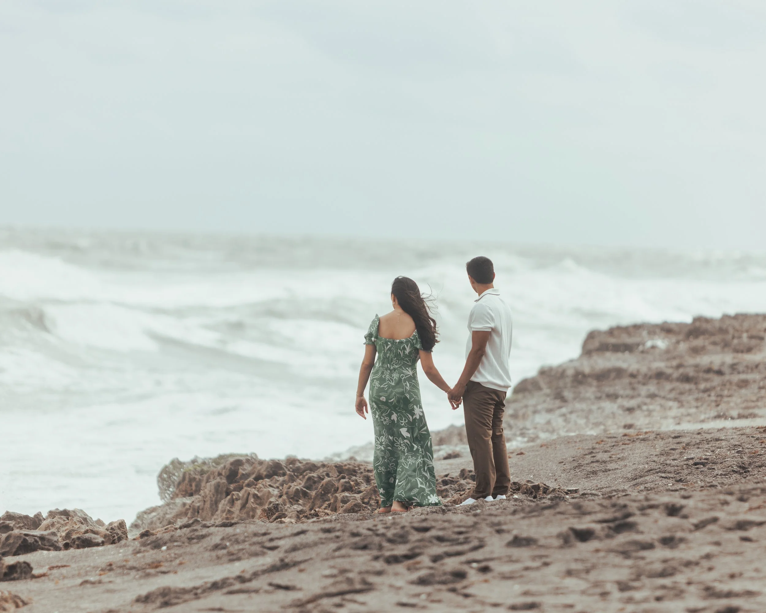 A couple holding hands walking on a rocky beach near the ocean, with waves crashing in the background.