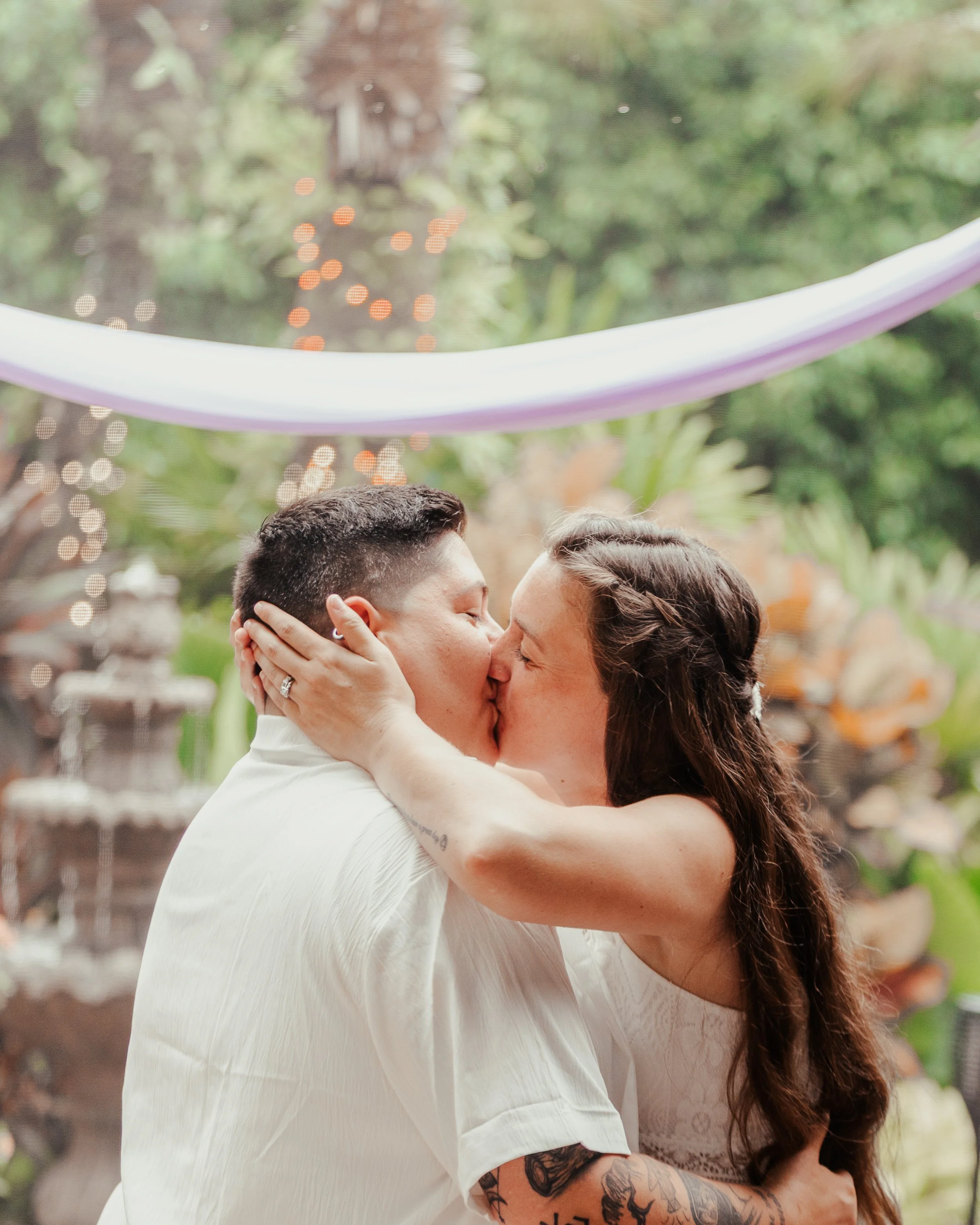 Two women kissing in an outdoor setting with greenery and string lights, one woman has long dark hair and tattoos, and the other has short dark hair.