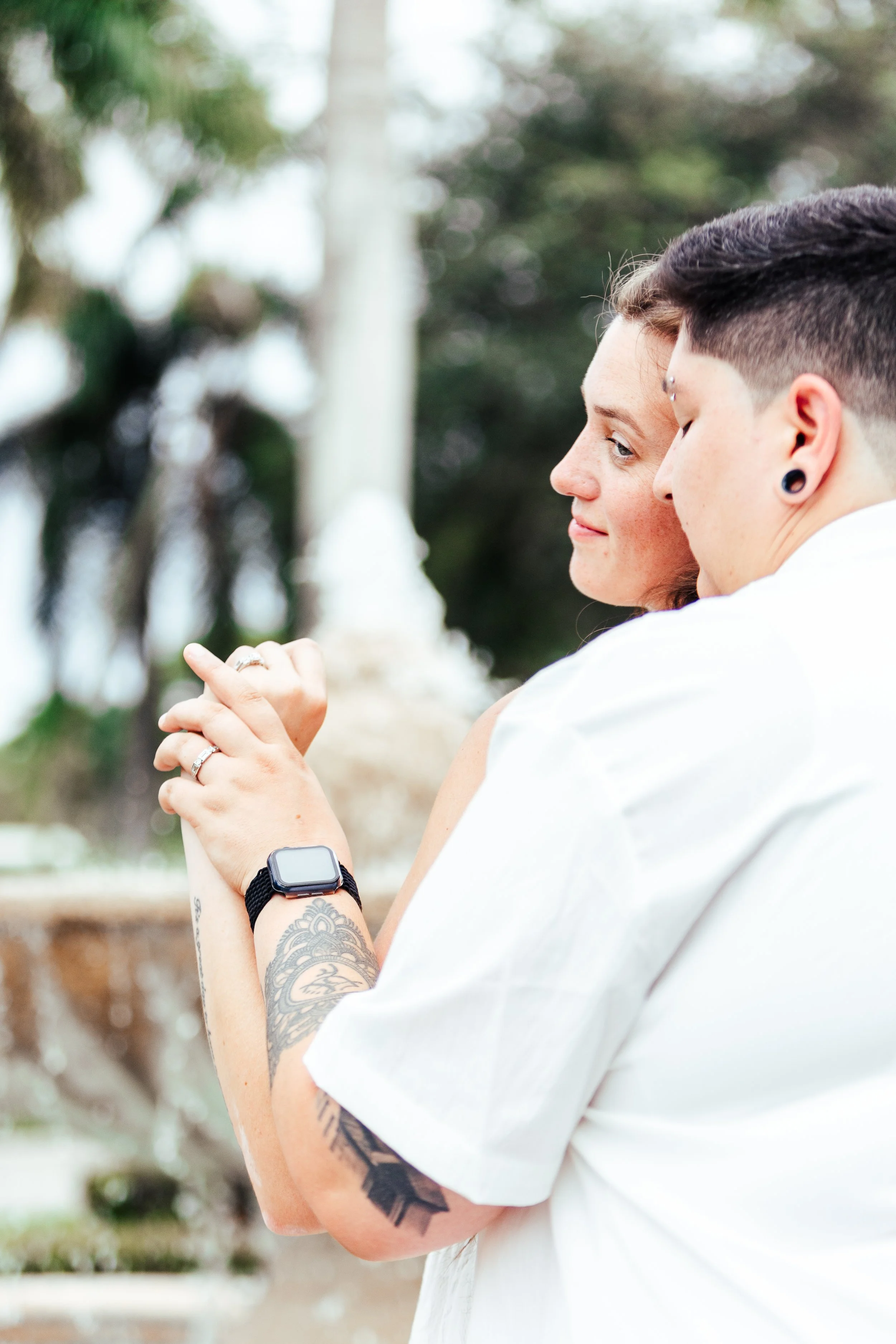 A couple with tattoos on their arms holding hands, with the woman wearing an Apple Watch, at an outdoor location with trees and a fountain in the background.