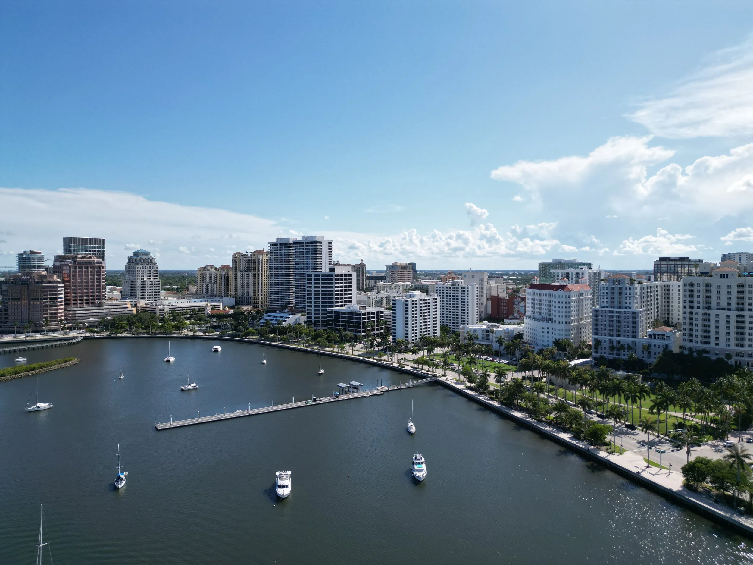Aerial view of a city skyline with high-rise buildings along a waterway with docked boats and a palm-lined sidewalk, under a partly cloudy sky.