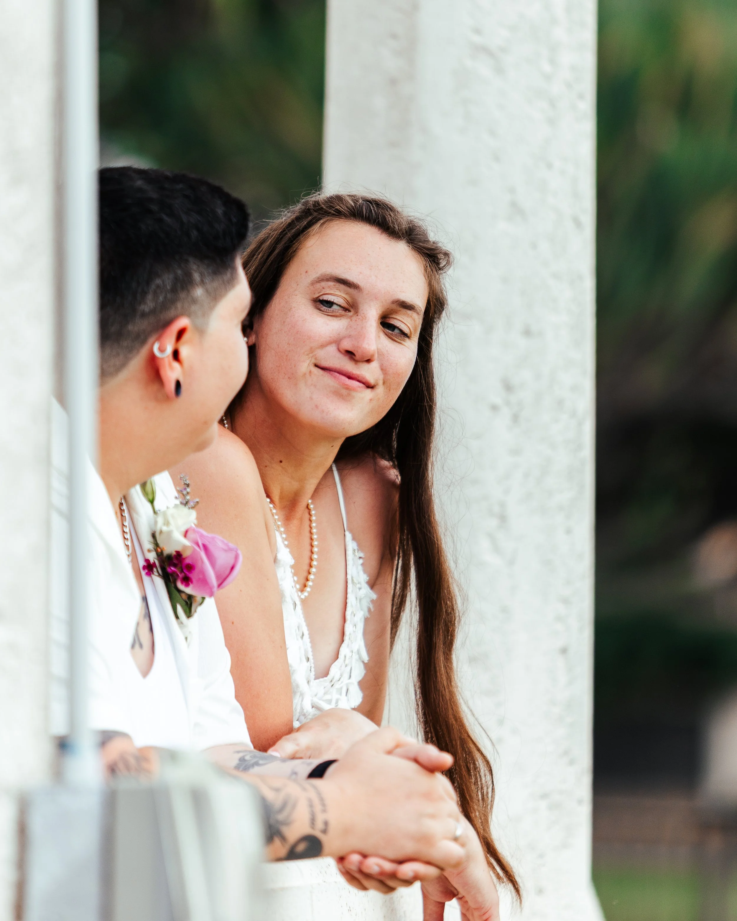 A woman looking at a person with short hair and tattoos, both dressed in wedding attire, leaning on a white railing.