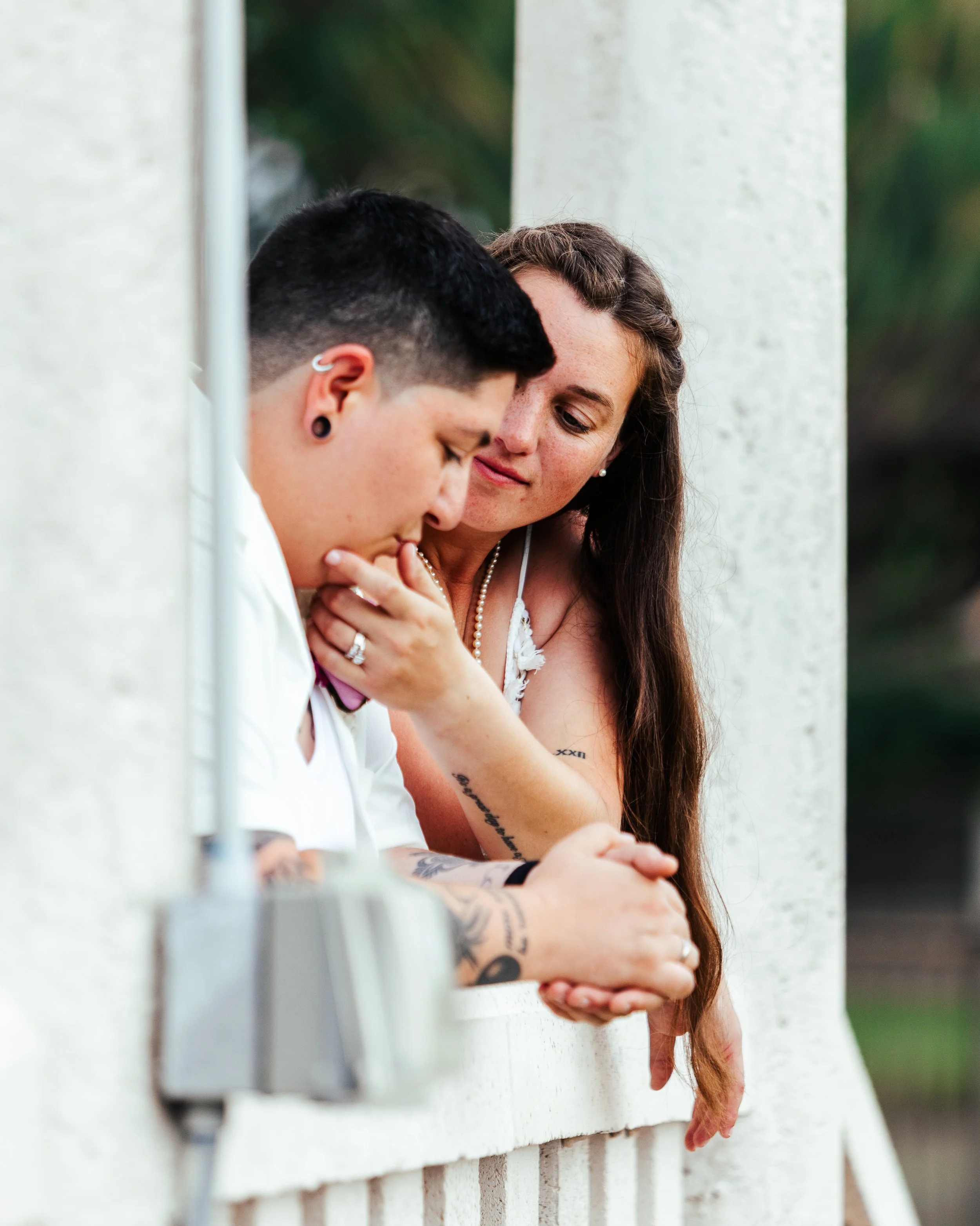 A person with short black hair and tattoos on their arm touching a ring on their finger, and a woman with long brown hair leaning on a white ledge, looking thoughtfully at the person.