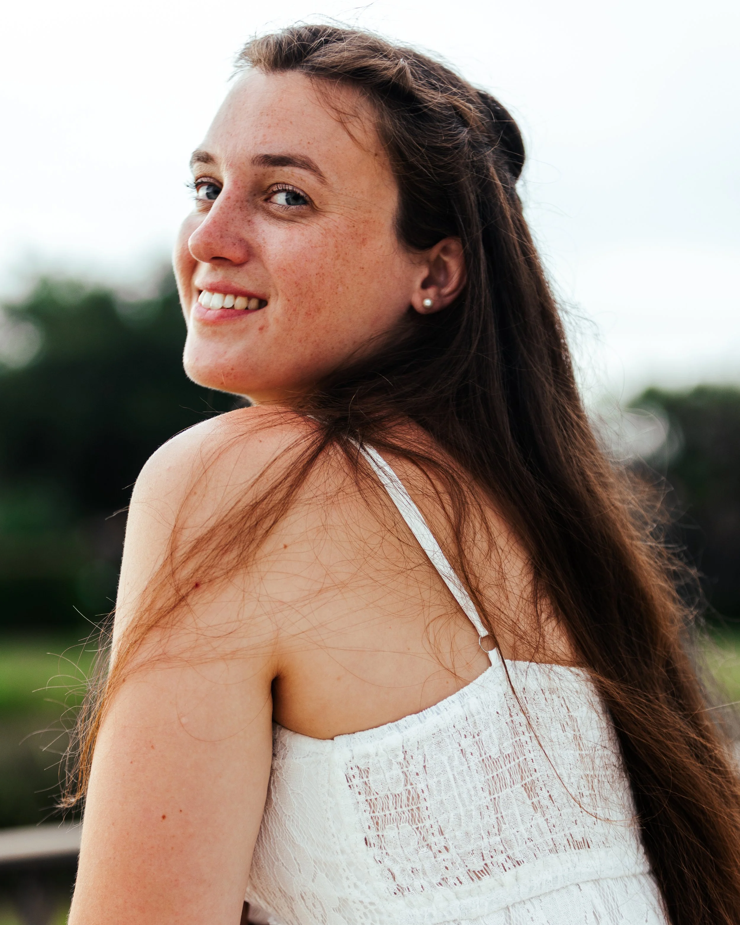 Close-up of a smiling young woman with long brown hair, wearing a white sleeveless dress, outdoors with blurred greenery in the background.