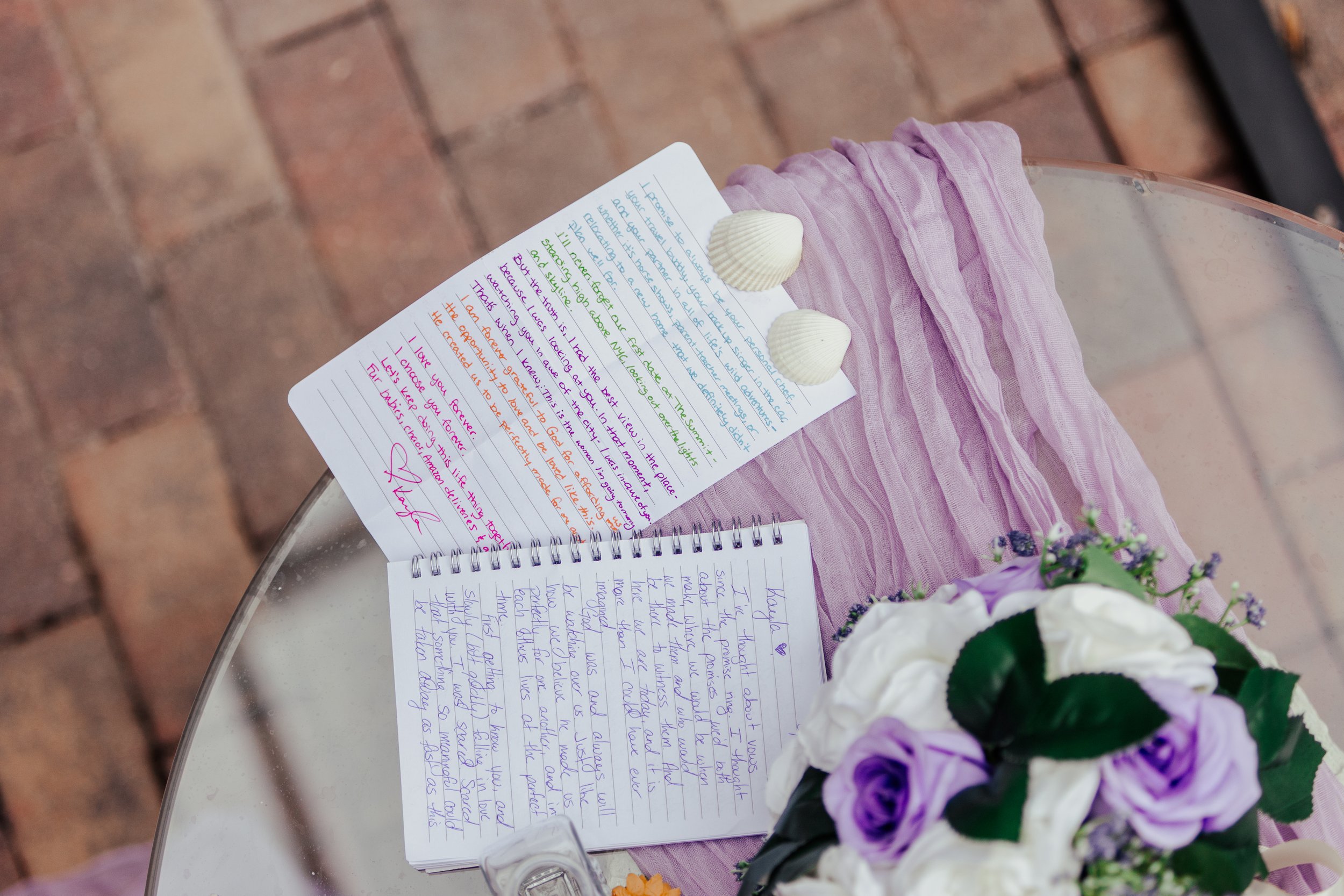 A printed letter and a handwritten note on a pink fabric, rose flowers, and seashells on a glass table with a brick ground background.