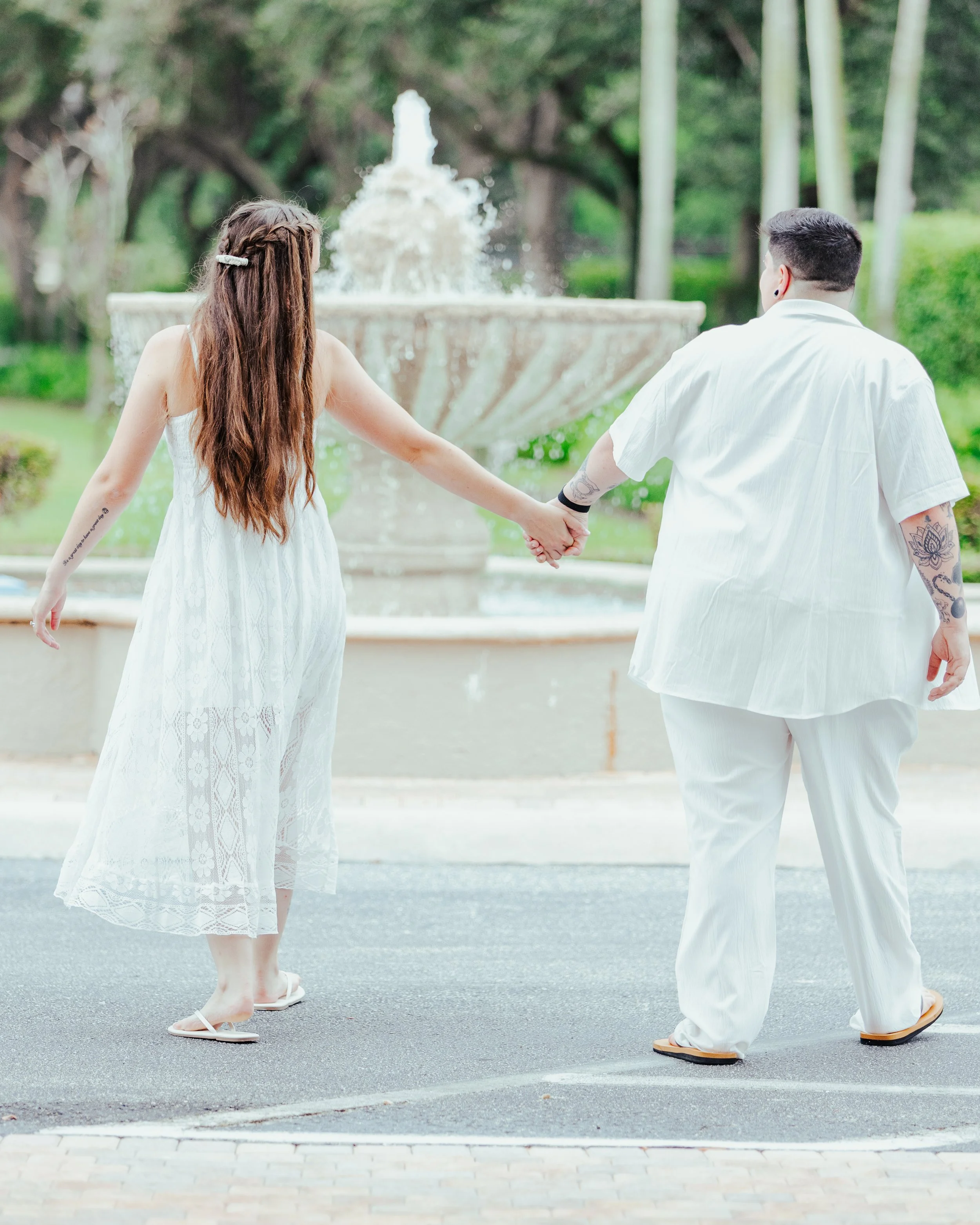 A couple holding hands while walking near a fountain in a park, both dressed in white.