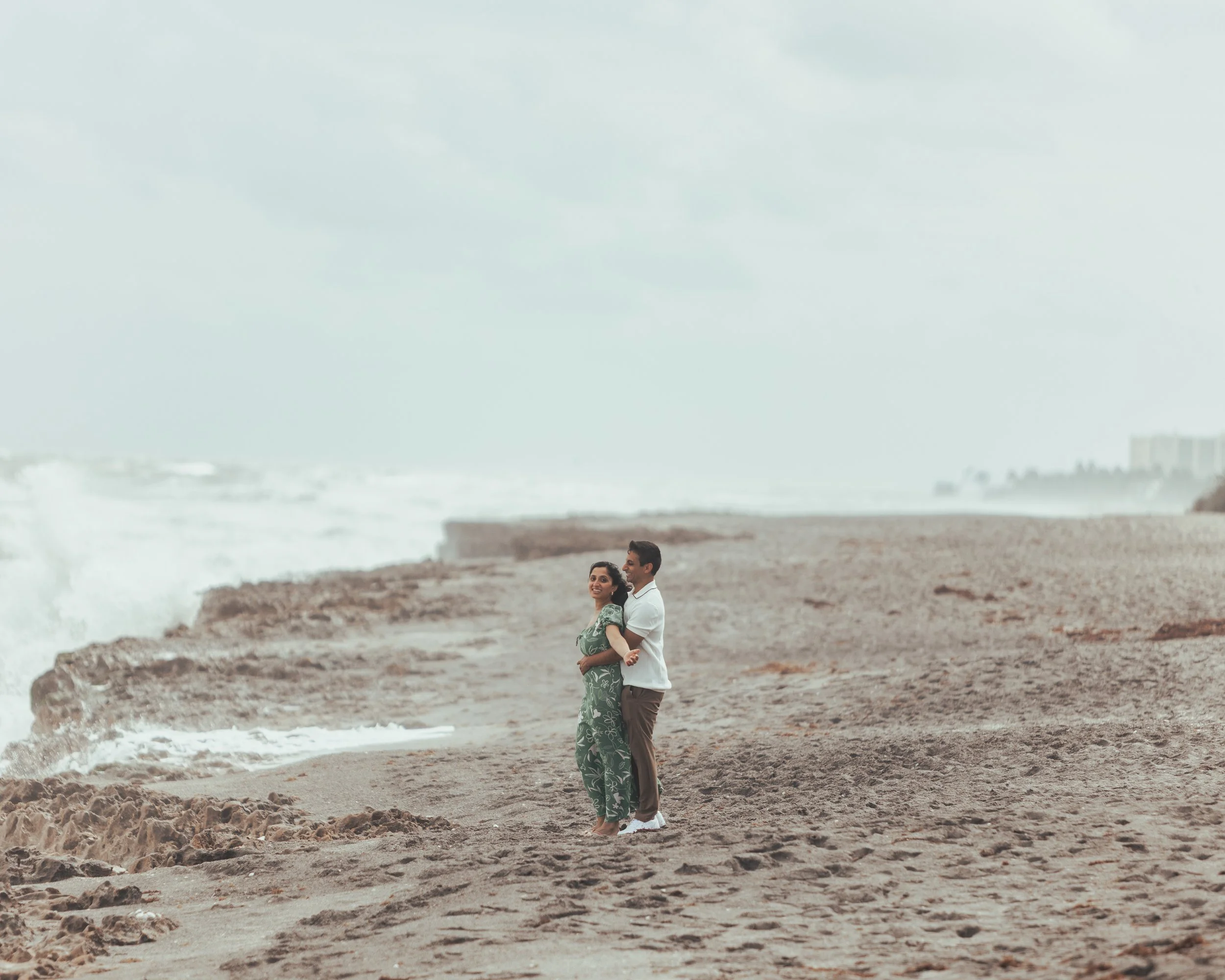 A couple stands on a rocky beach near the ocean, embracing each other, with waves crashing in the background and a cloudy sky overhead.