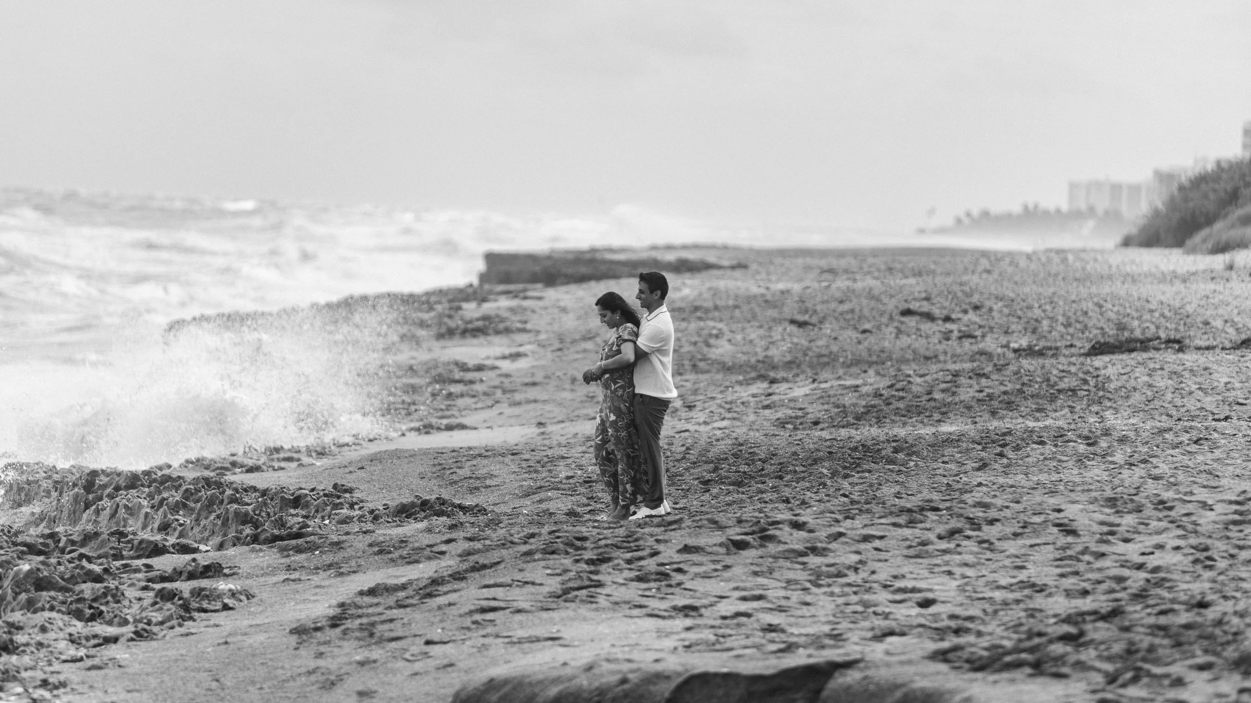 A black and white photo of a couple standing on a beach, embracing and looking out at the ocean, with distant city buildings in the background.