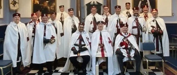 Group of men in ceremonial robes and hats, standing and seated in a formal room with framed portraits, chandeliers, and chessboard-patterned carpet.