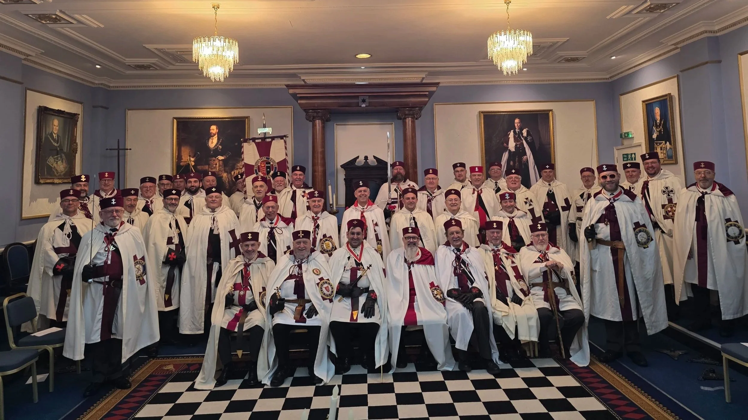Group of men in ceremonial robes and hats, standing and seated in a formal room with framed portraits, chandeliers, and chessboard-patterned carpet.