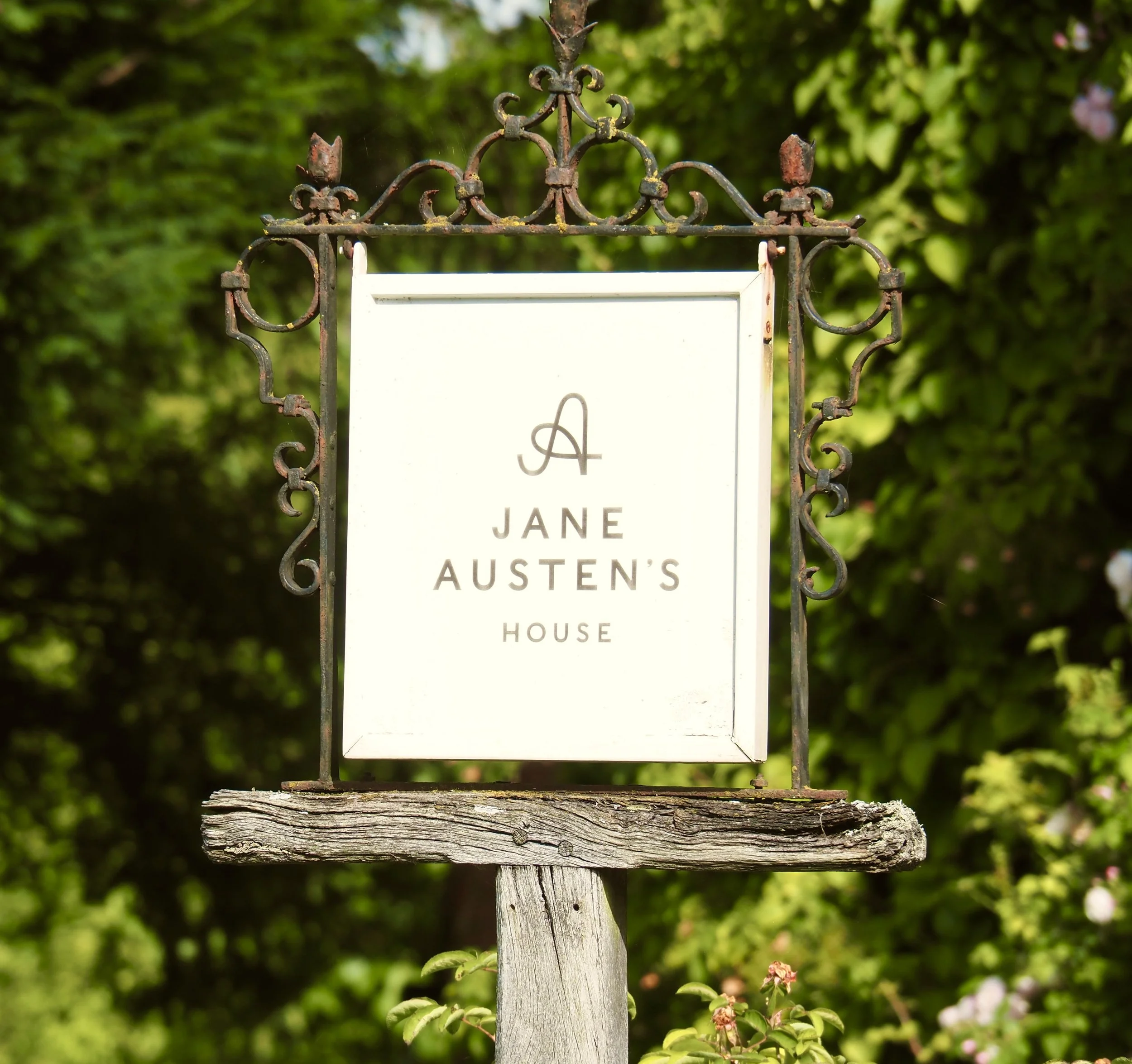 Sign for Jane Austen's House with decorative iron frame and wooden post, set outdoors with green foliage in the background.