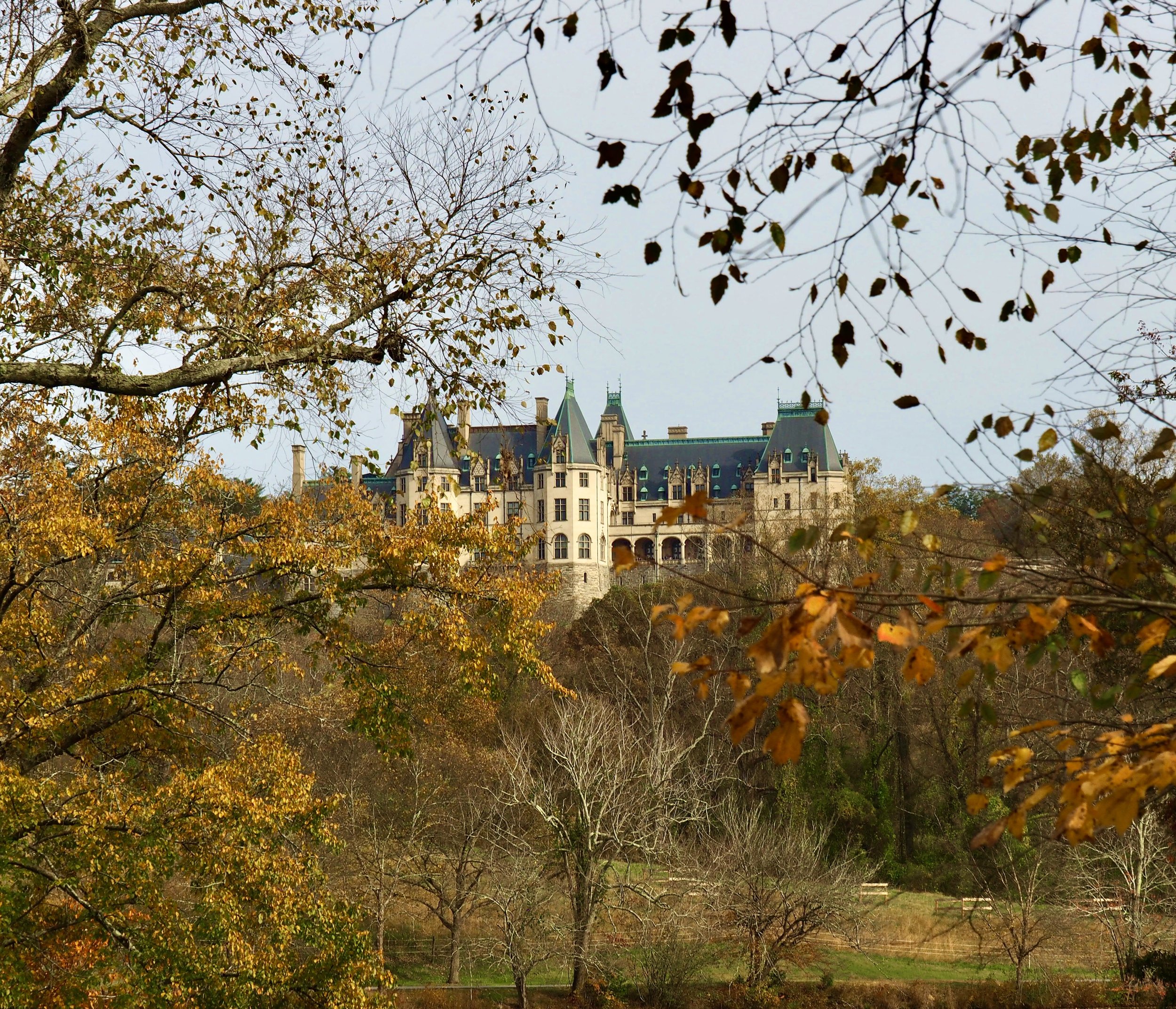 A castle surrounded by trees with autumn-colored leaves, situated on a hill beneath a cloudy sky.