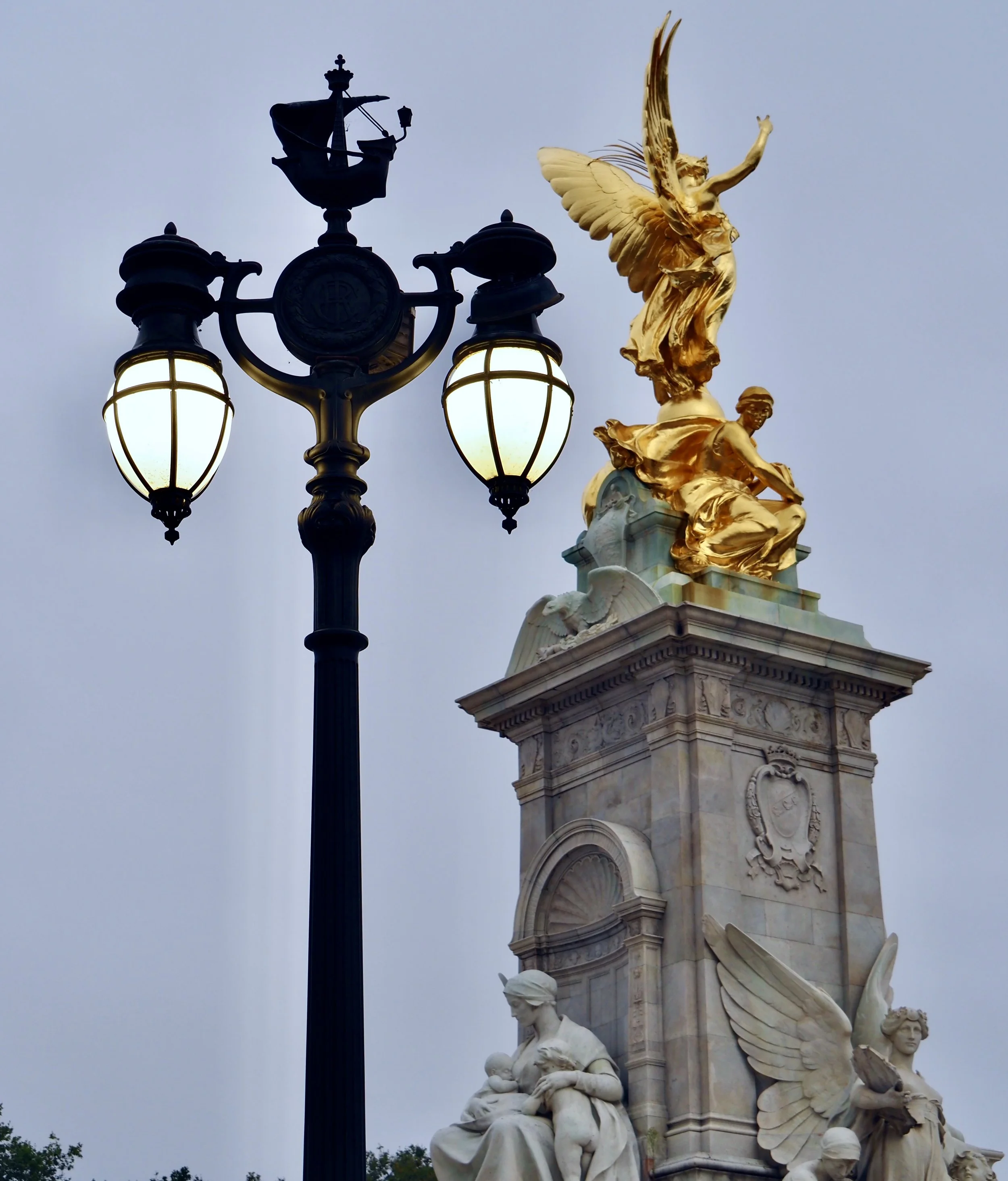 A decorative street lamp with four glowing bulbs and a tall monument featuring gilded statues and sculptures, including a winged figure at the top and seated allegorical figures at the base, set against a cloudy sky.