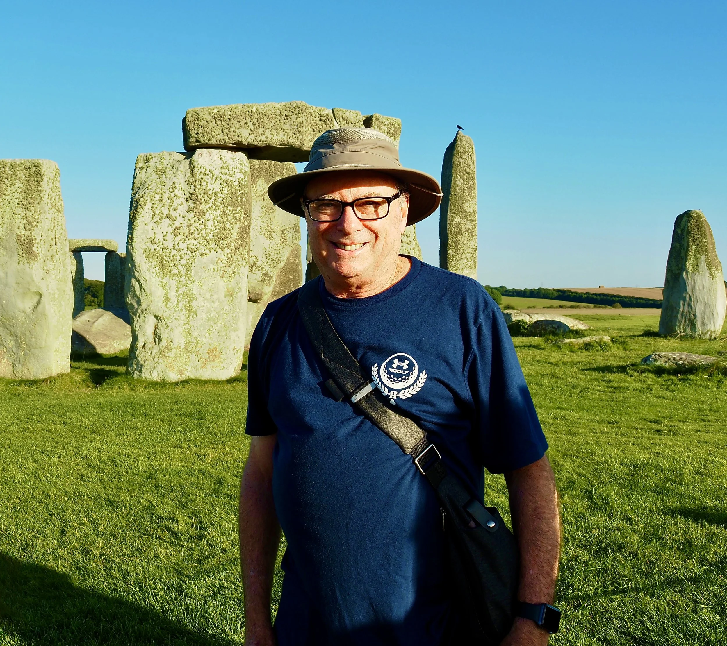 A smiling man wearing glasses, a tan hat, a navy blue T-shirt, and a black shoulder bag stands in front of Stonehenge on a sunny day.
