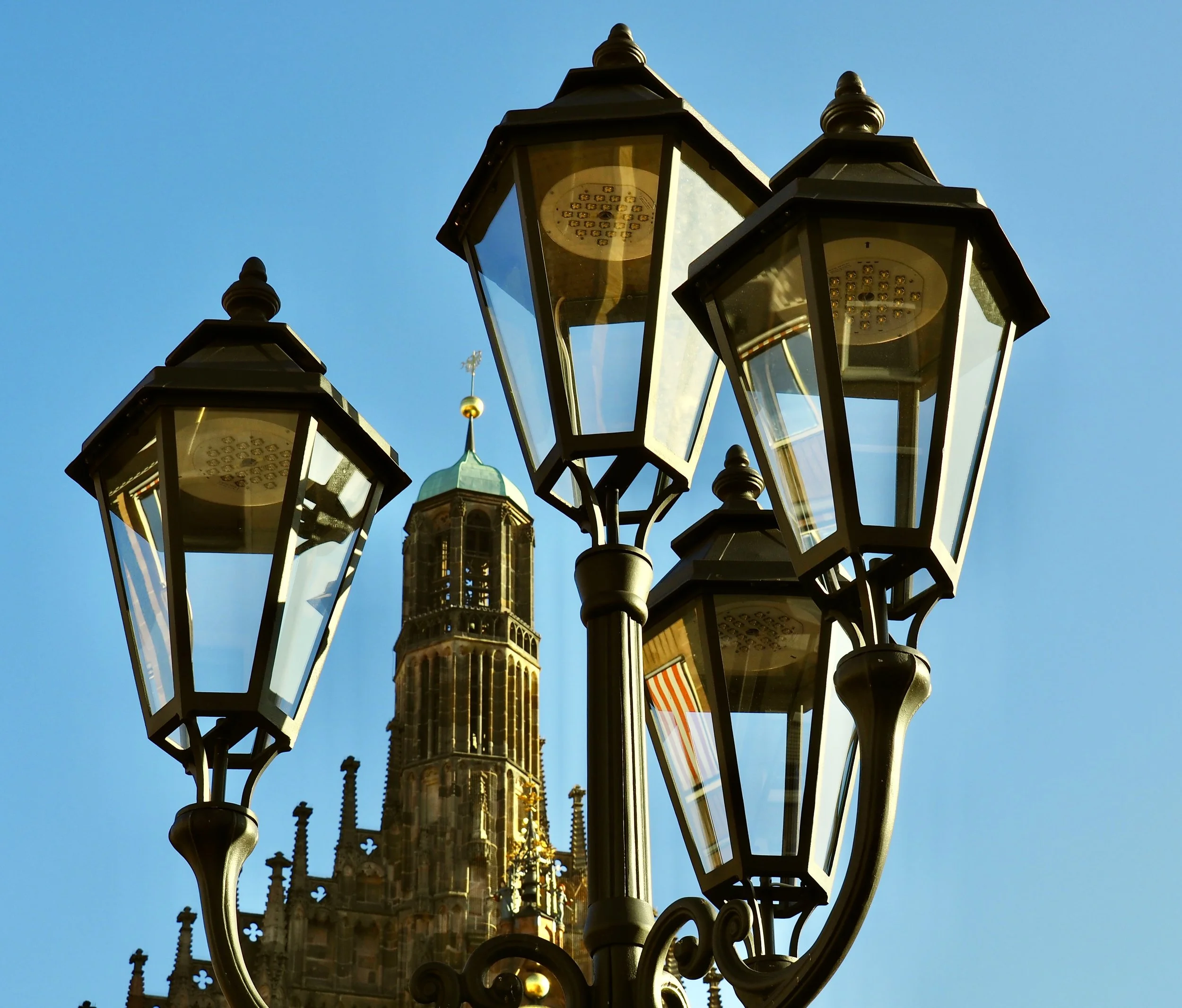 A close-up view of five ornate black street lamps with glass covers, set against a bright blue sky. In the background, a historic building with a tall, decorative green and gold spire is visible.