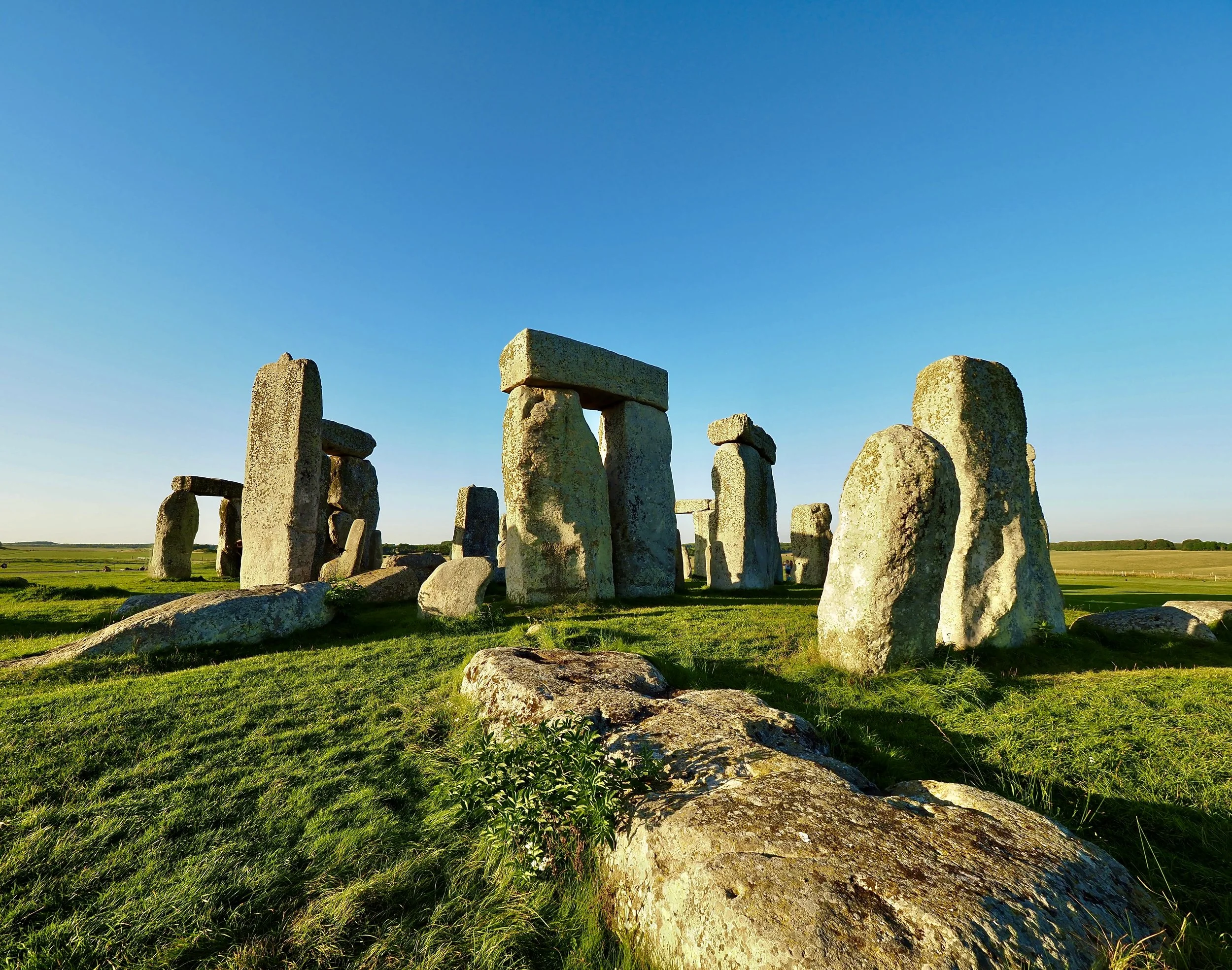 Sunlit view of Stonehenge, a prehistoric stone circle monument, with large upright stones and some horizontal stones forming a ring, set in a grassy plain under a clear blue sky.