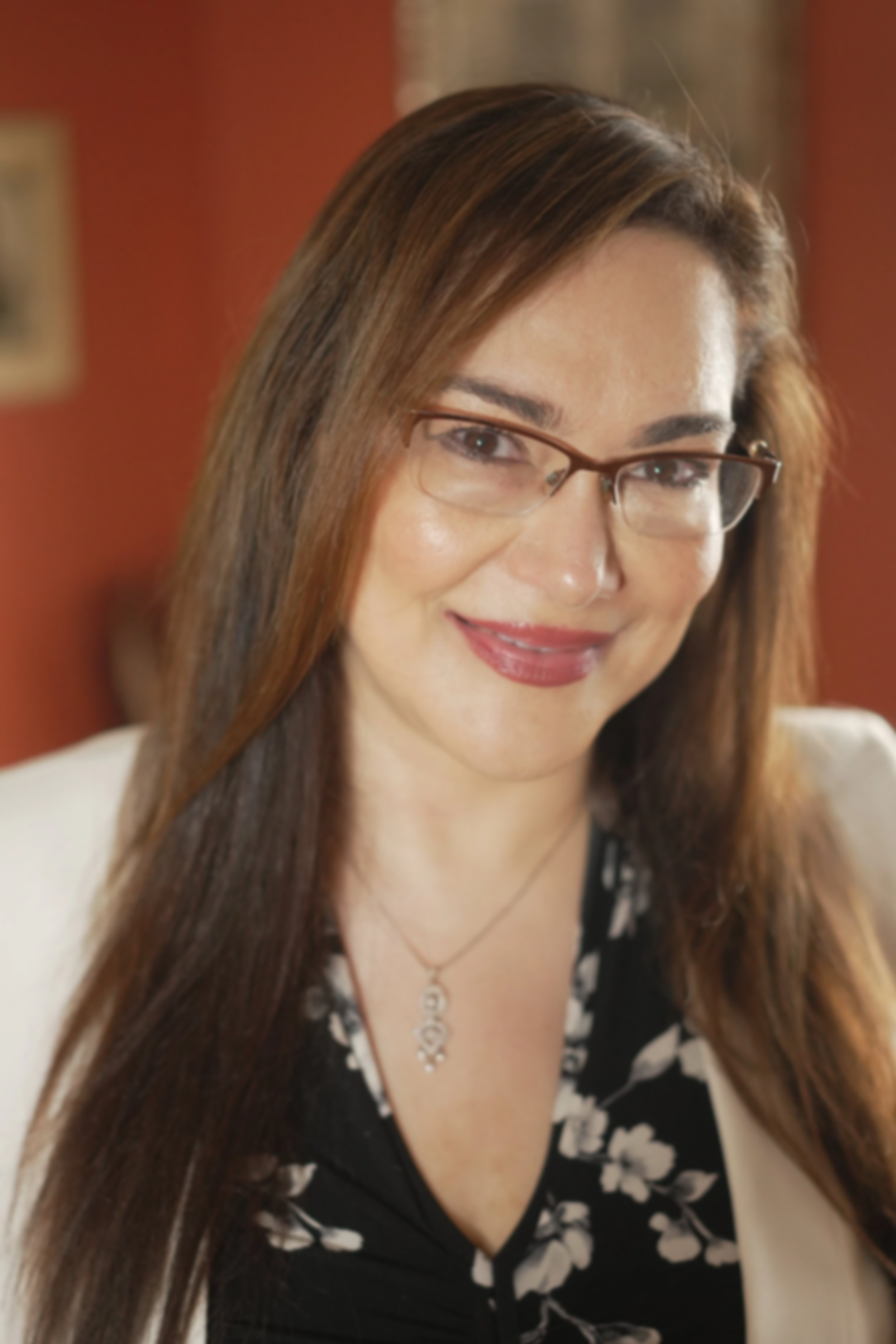 A woman with long brown hair, glasses, and a silver necklace, smiling at the camera, standing indoors with a reddish wall in the background.