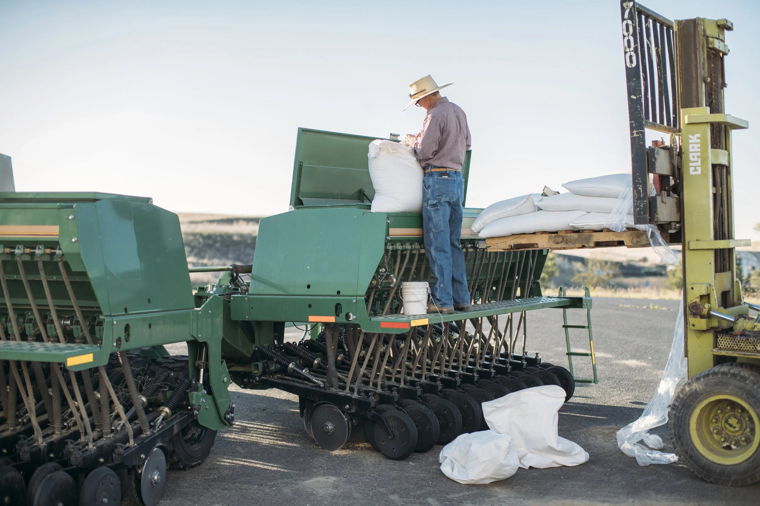 A farmer stands on a combine, sealing a bag of wheat. There are rolling hills of wheat in the blurred background.