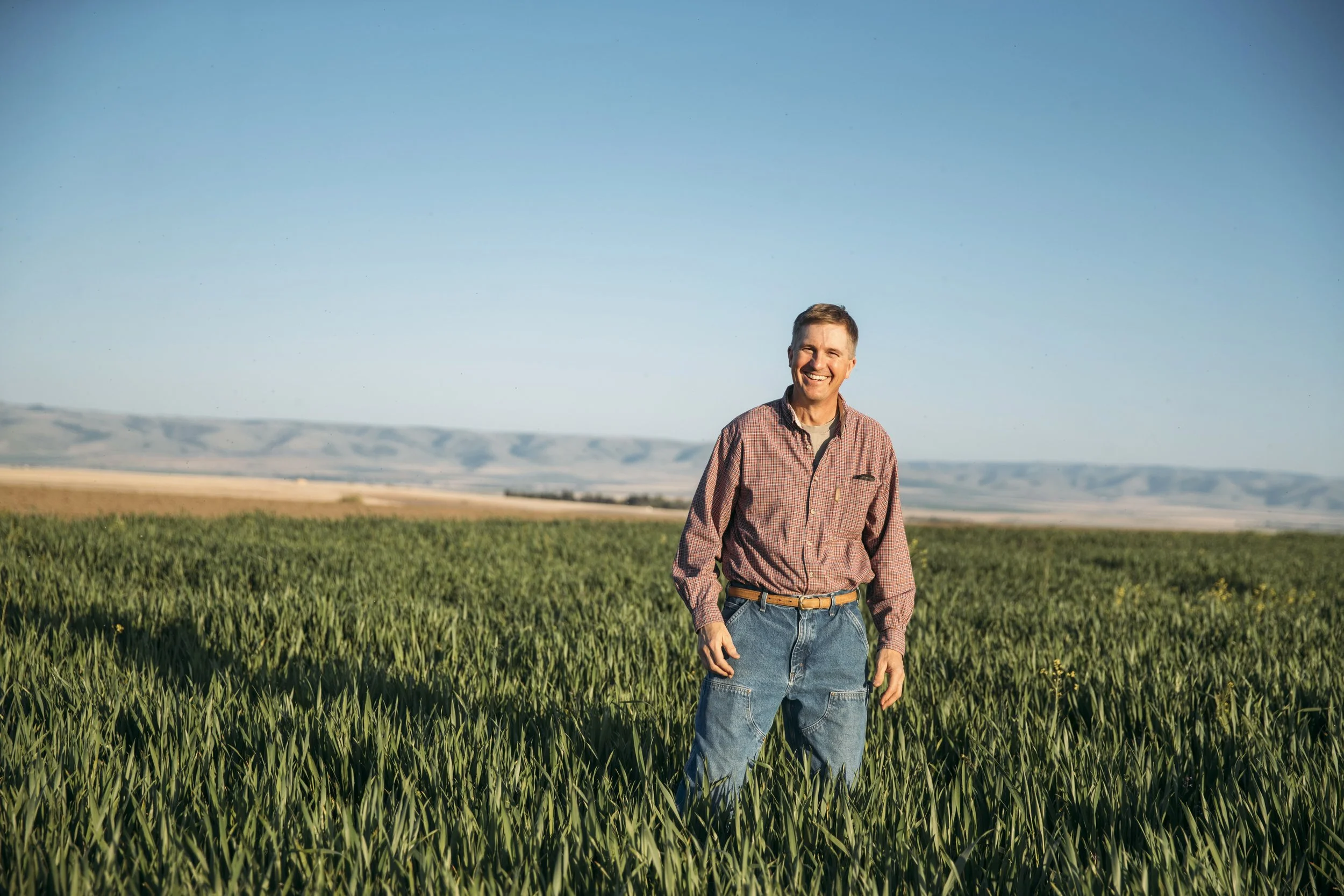 Eric Nelson stands in a field of green crops. He wears a light red, patterned shirt and jean pants.