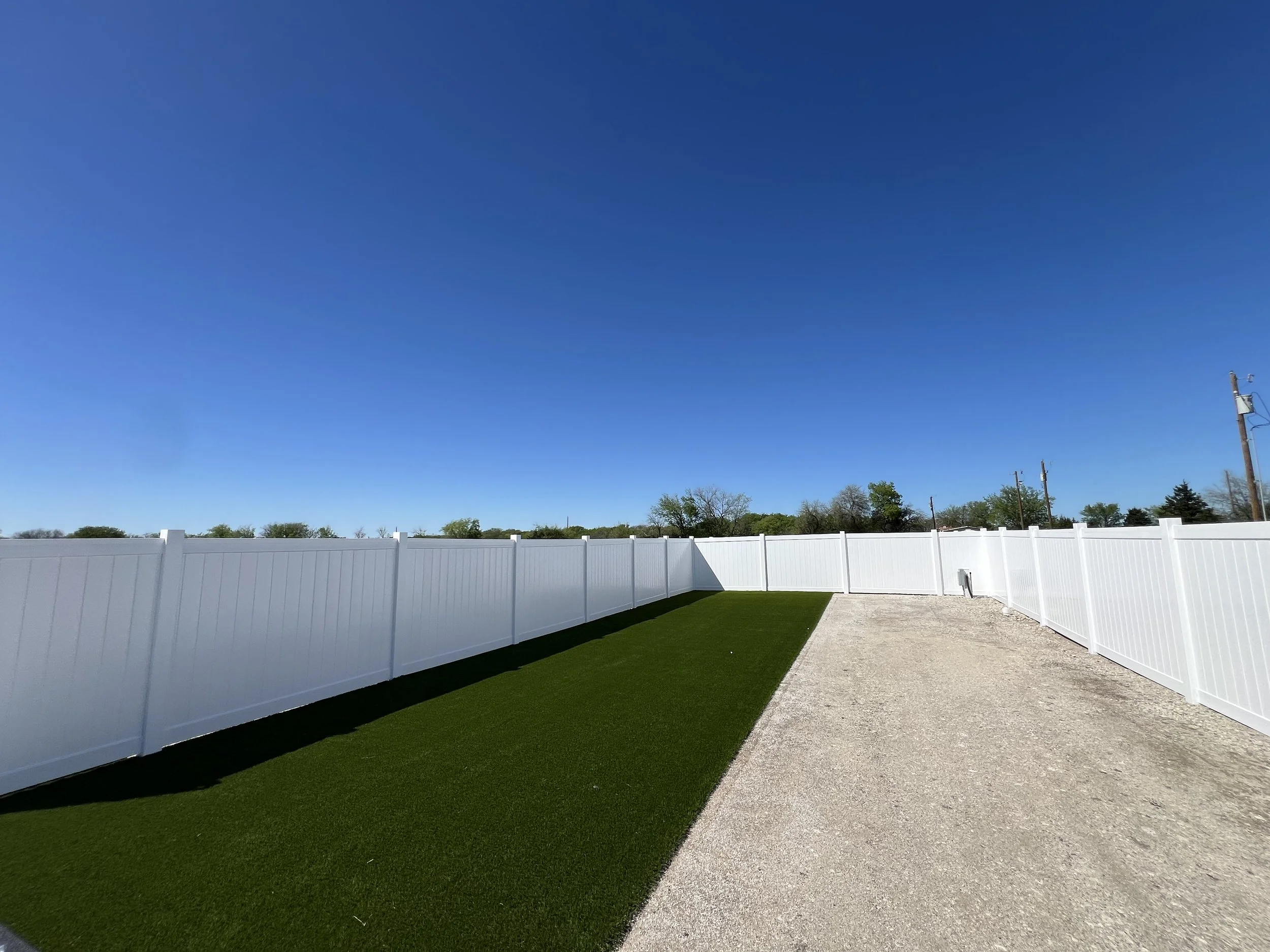 Fenced RV lot with turf strip, gravel path, and trees in the distance.

