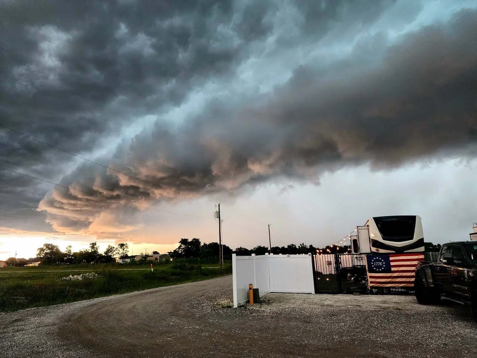 Dark, ominous storm clouds loom over a rural area during sunset, with a row of trees and houses in the distance. In the foreground, there is a gravel lot with a white fence, a large RV decorated with the American flag and the year 1776, and a black t