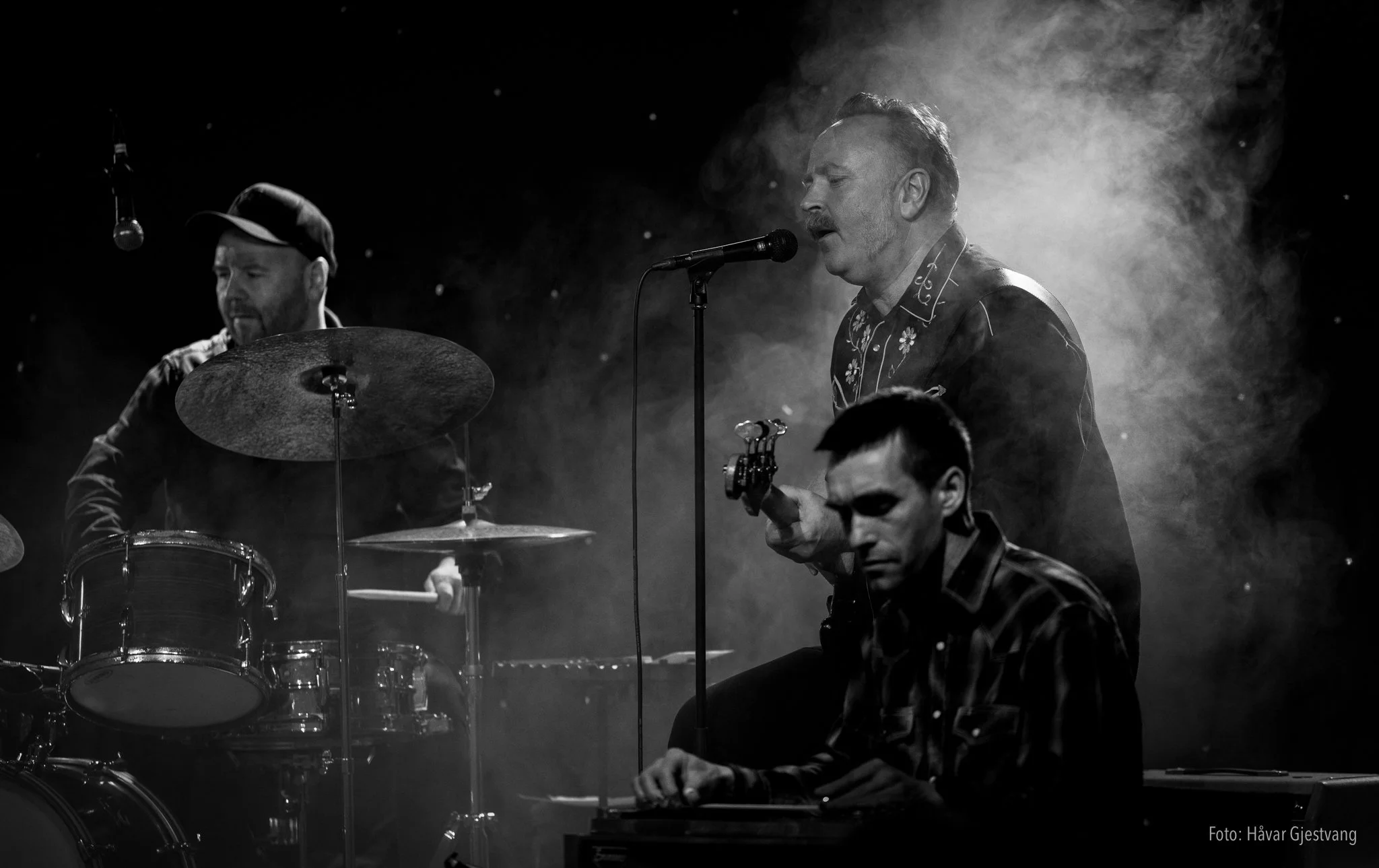To menn som spiller musikk på en scen, en med trommer og den andre med en mikrofon og gitar. Bildet er i svart-hvitt.