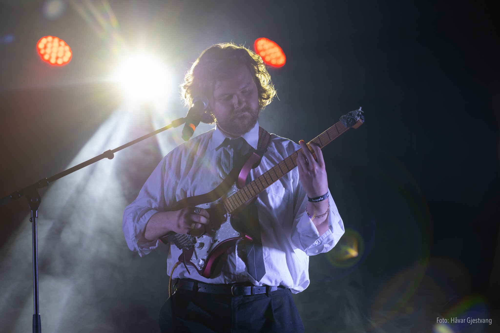 En mann med skjegg spiller elektrisk gitar under en konsert, med stort lys bak ham og røde lyspærer som lyser opp scenen.