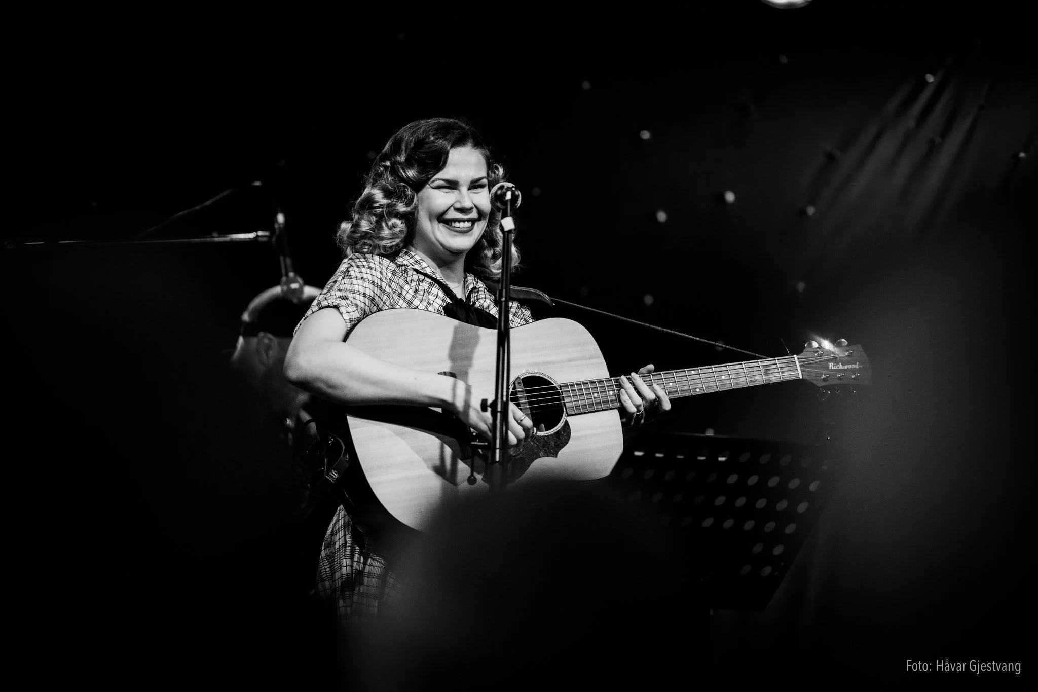 Frida. En kvinne med krøllet hår spiller akustisk gitar og smiler på en scenes konserten. Bildet er i svart-hvitt.