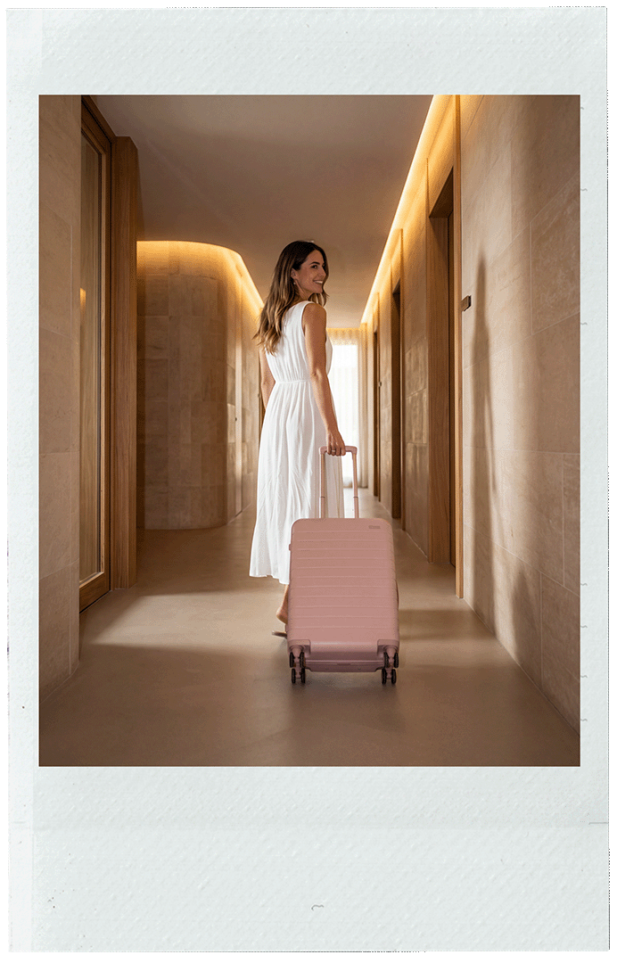 A woman in a white dress pulling a pink rolling suitcase down a modern, well-lit hallway with wooden accents.