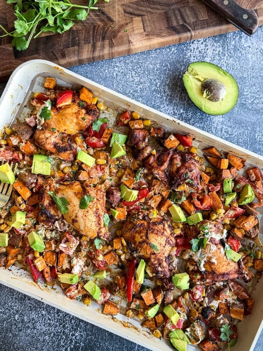 chipotle chicken with roasted veggies on a sheet pan with half an avocado next to the pan and some fresh cilantro on a wooden cutting board