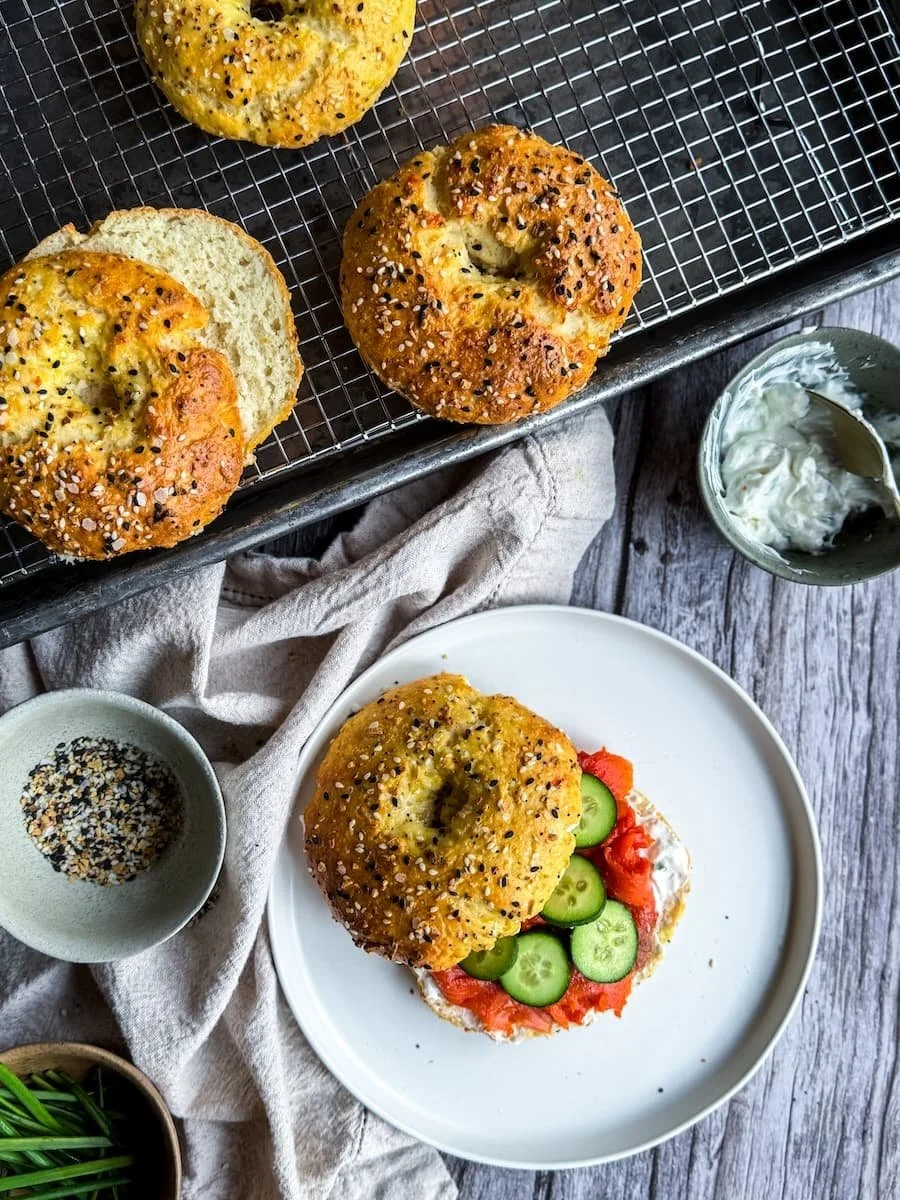 overhead shot of savory protein bagels - 3 are on a wire rack and one is on a plate with cream cheese, cucumbers, and lox