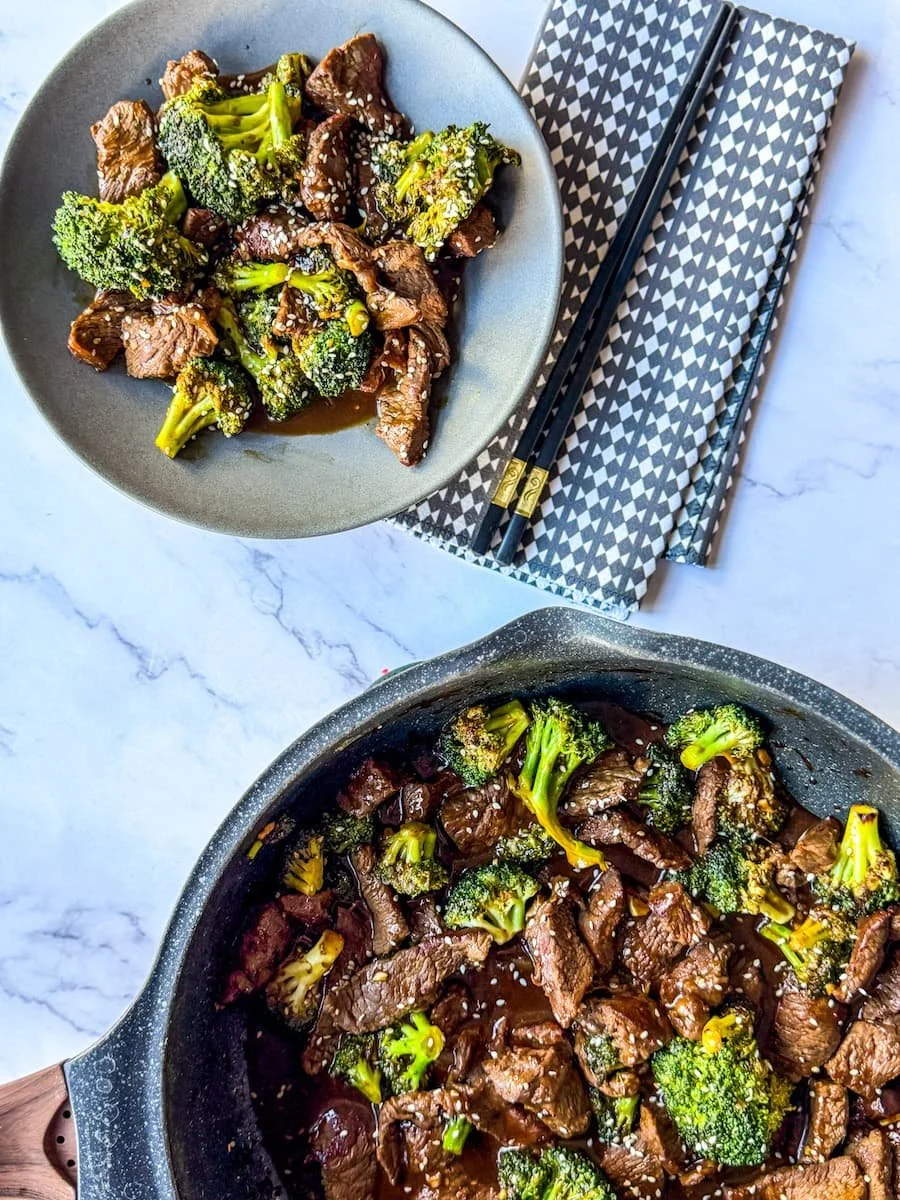 overhead shot of beef broccoli recipe with a portion on a plate next to chopsticks and a cloth napkin and the rest in the skillet