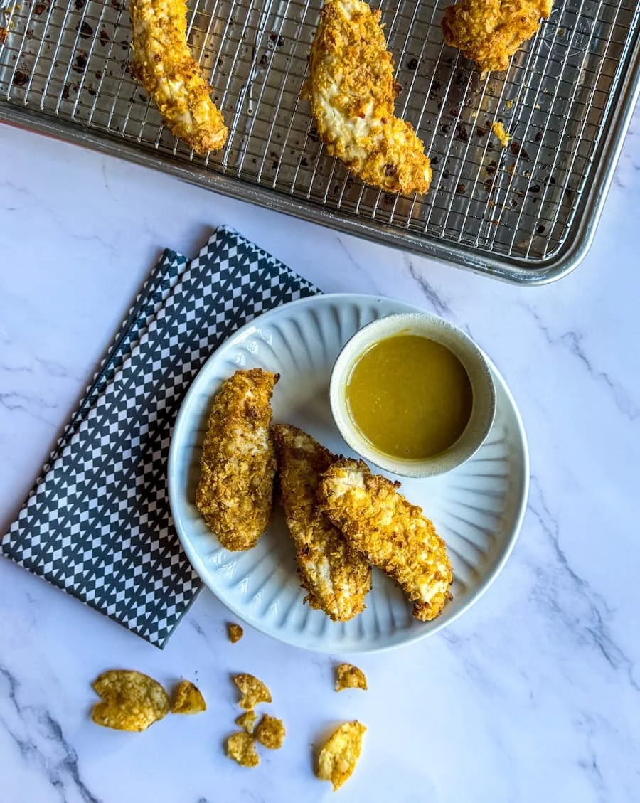 baked chicken tenders - three tenders on a plate with a dish of honey mustard sauce next to it and a pan with additional chicken tenders in the background