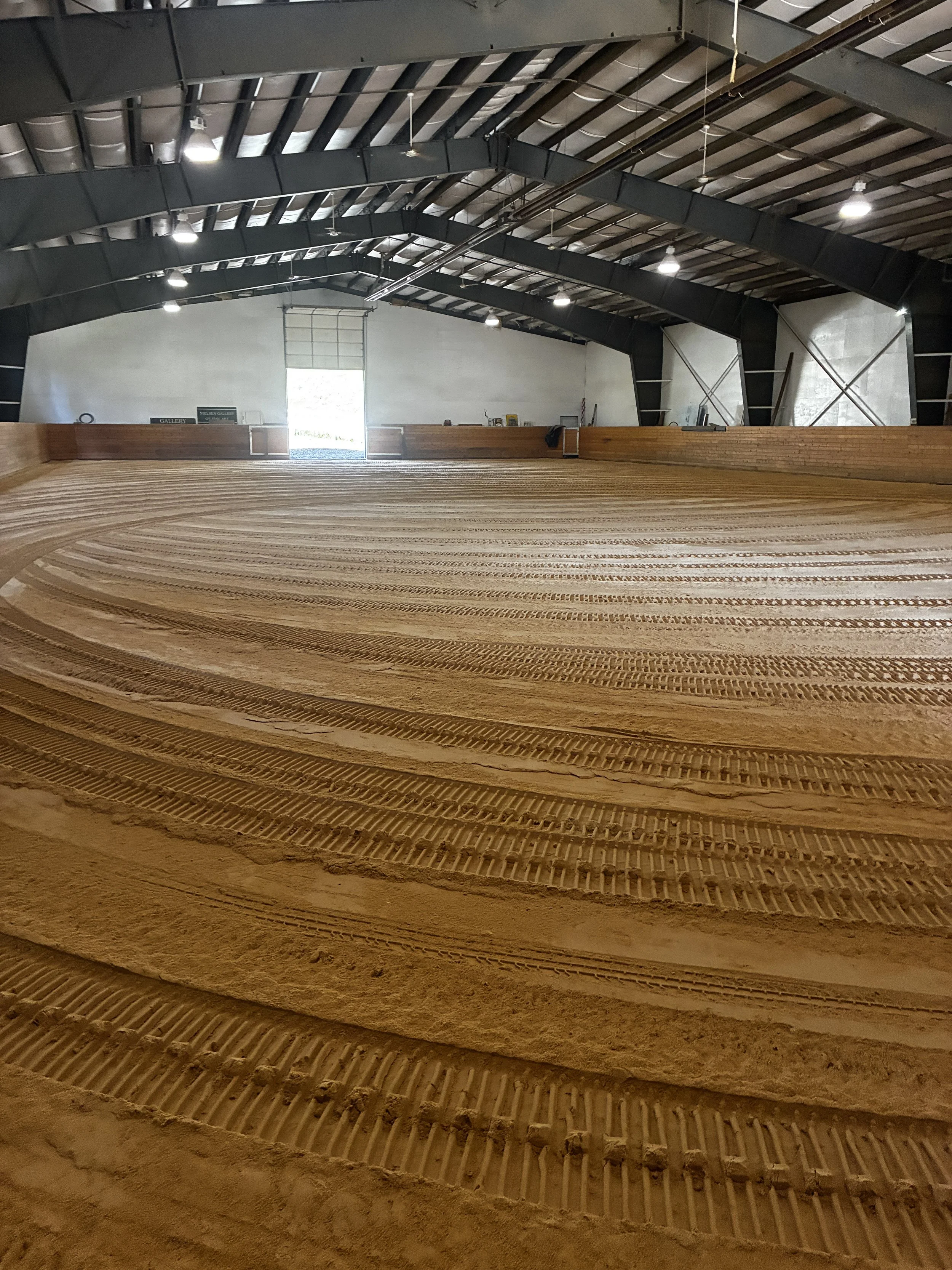 An indoor arena with a dirt riding surface, showing lift tracks in the dirt for a horse riding ring, with a metal roof and open roller door at the far end.