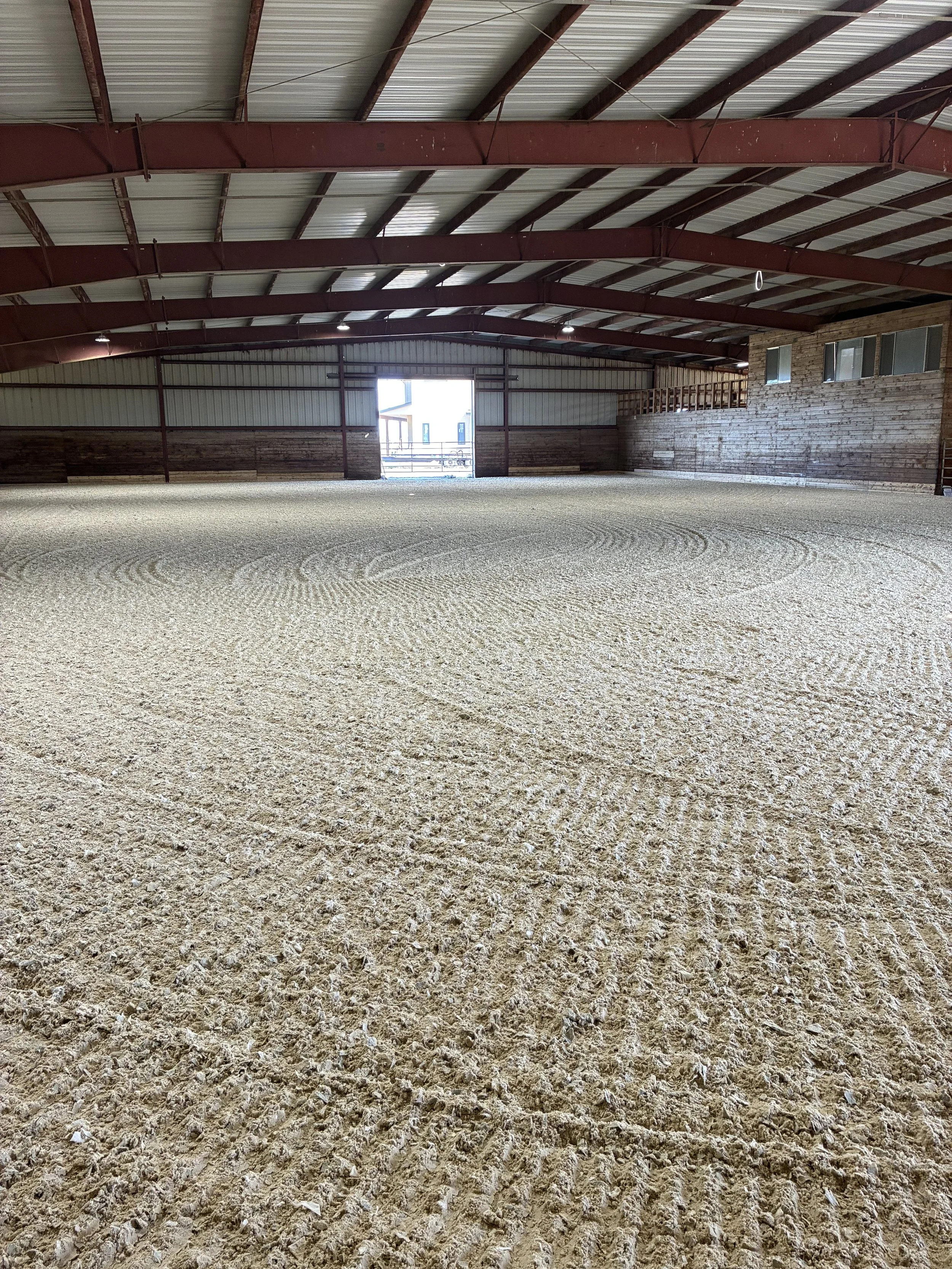 An empty indoor riding arena with a dirt or sand surface, wooden and metal structures, and a large door open to the outside.