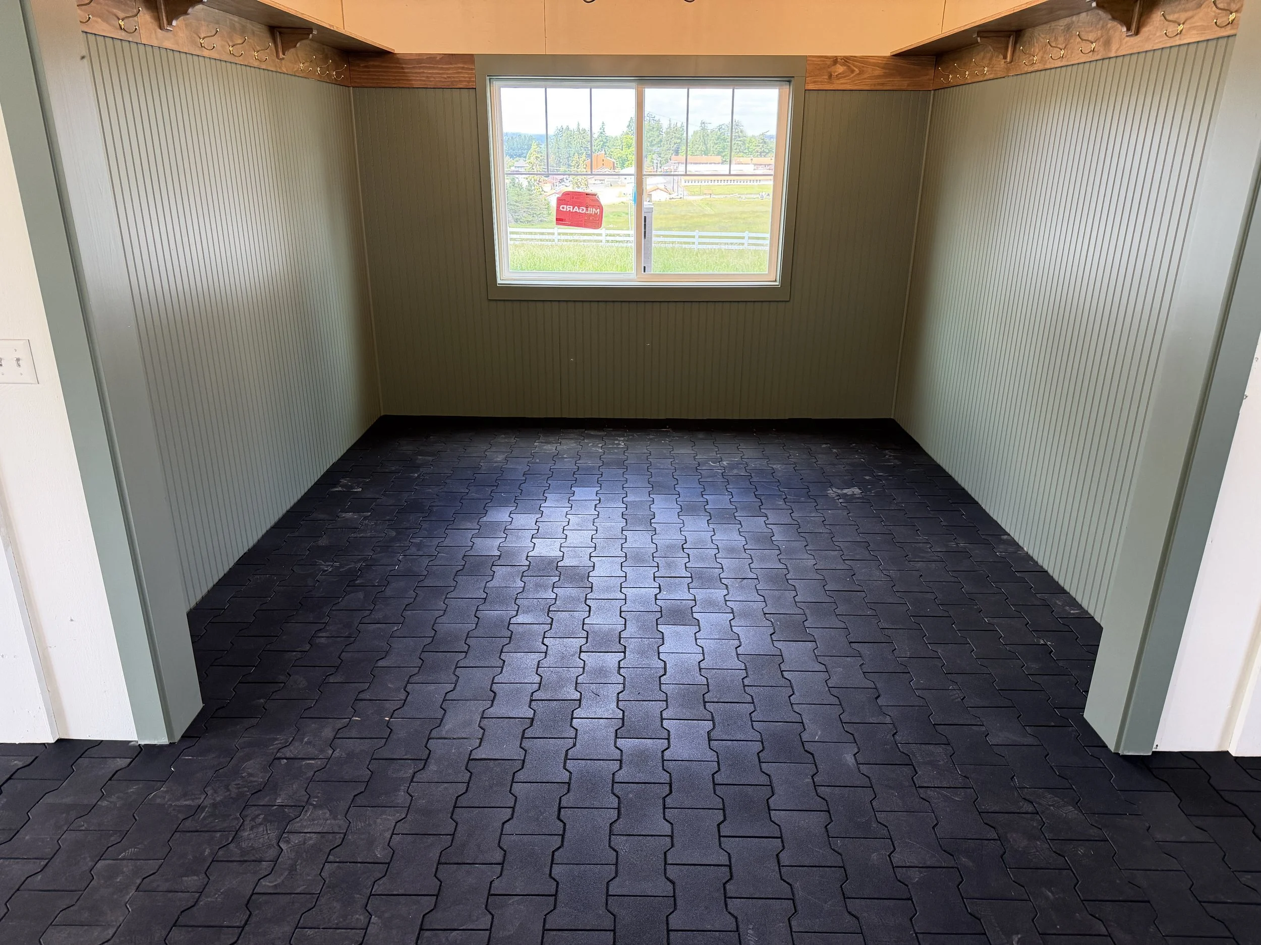 Empty room with black rubber interlocking floor tiles, beige panel walls, two large windows showing outdoors with green grass, trees, and a red sign on one window. Wooden hooks and coat rack above the windows.