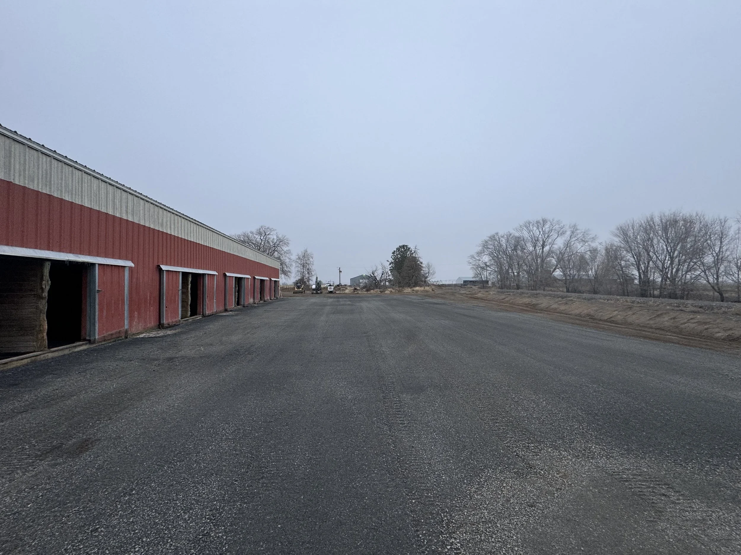 A red metal barn with large open doors on a gravel lot, located in a rural area with leafless trees and overcast sky in the background.