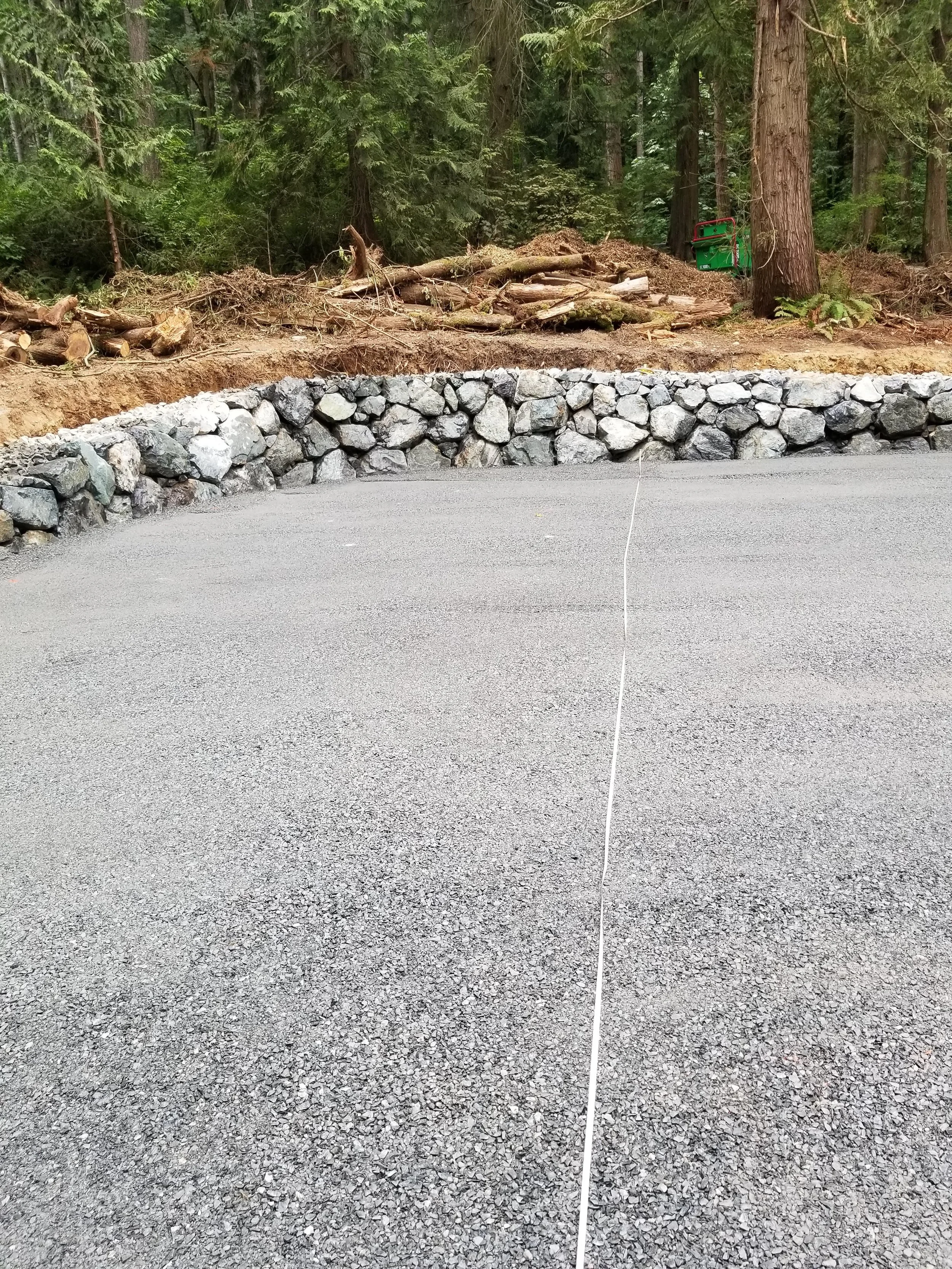 A gravel road in front of a stone retaining wall, with a dirt and forested area behind. There is a small green equipment box in the background near a tree.