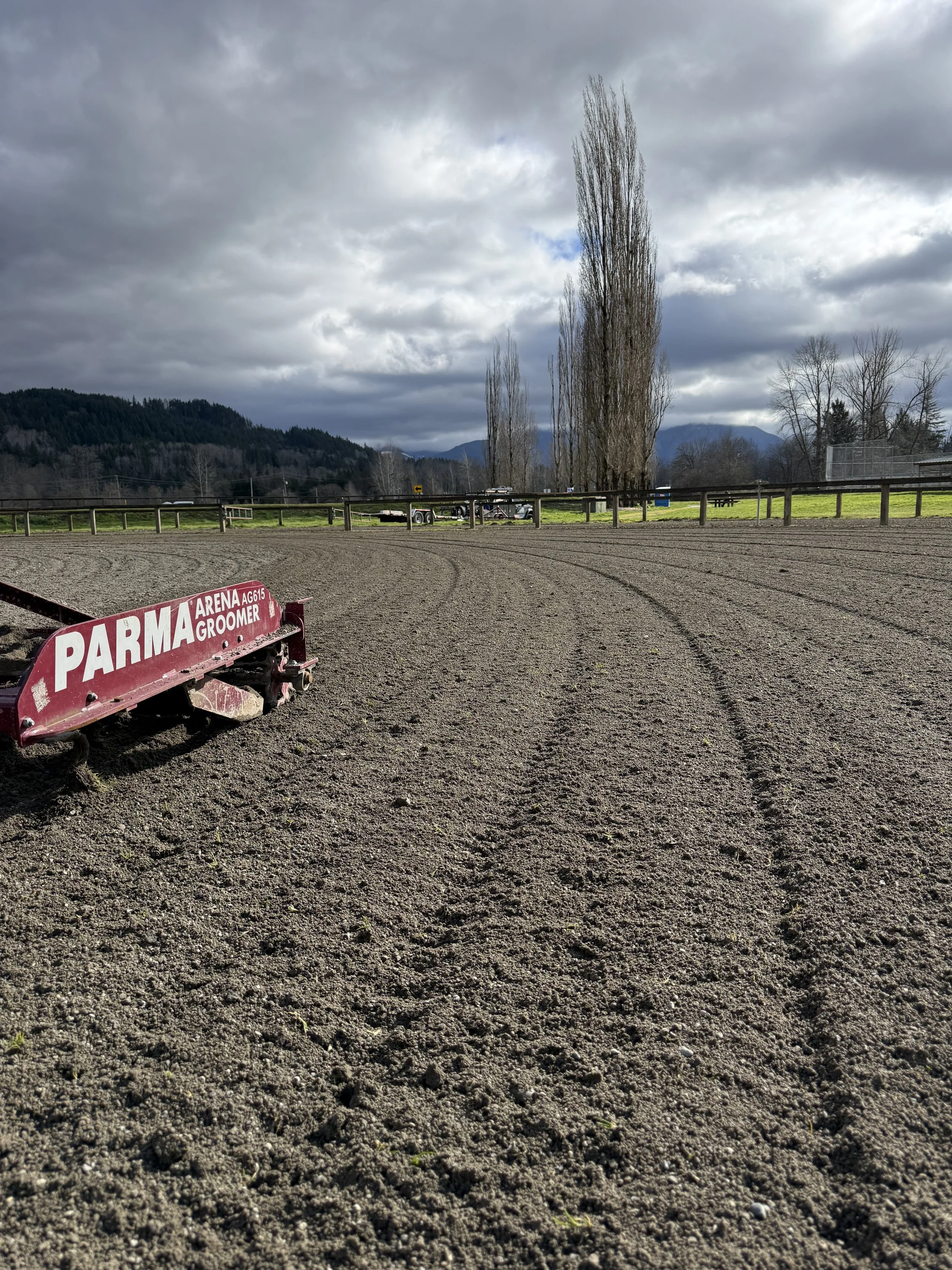 View of a dirt racetrack with a red horse grooming equipment in the foreground, a fence, trees, hills, and cloudy sky in the background.