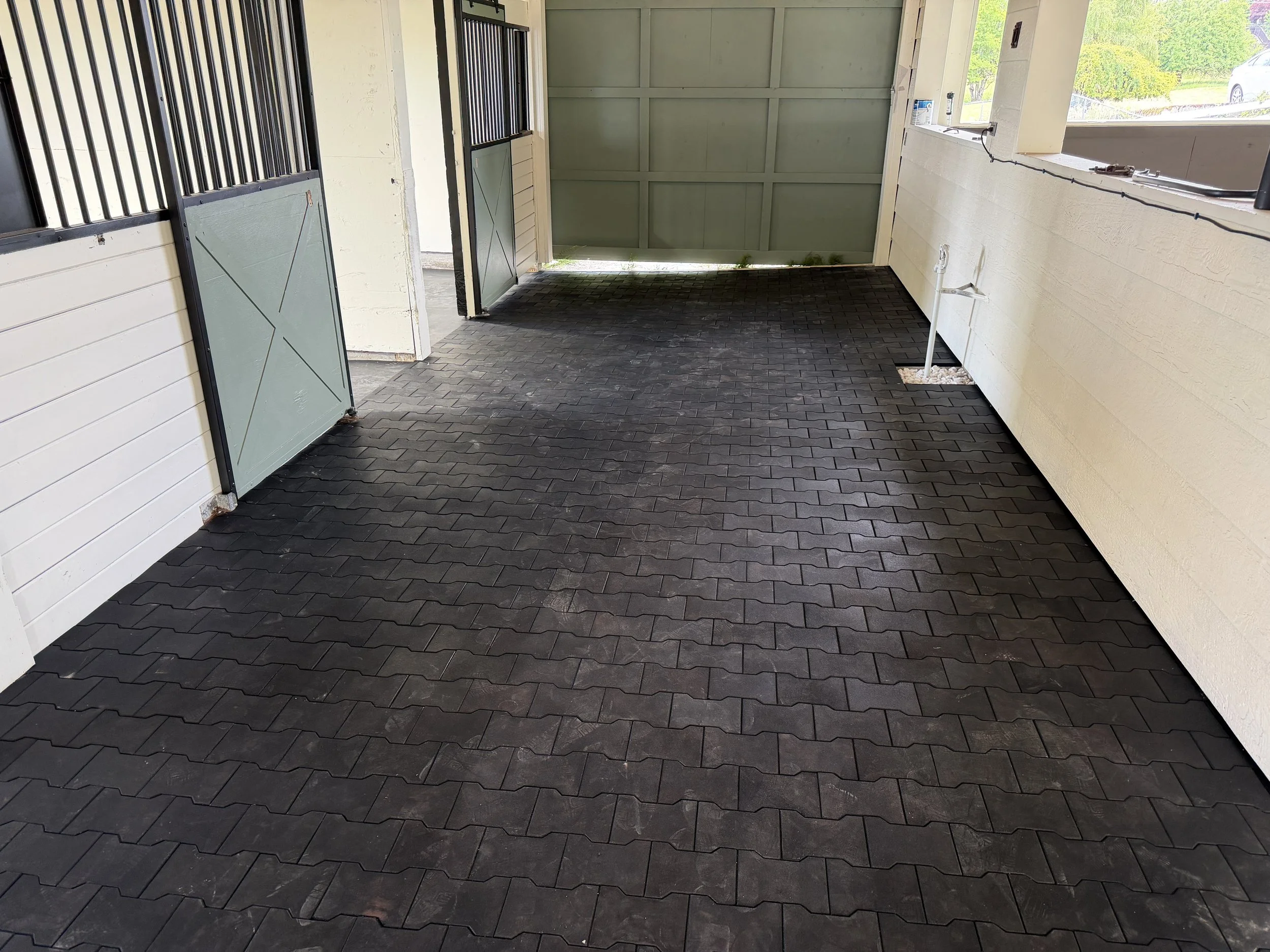 A garage or covered outdoor area with black brick paver flooring, light-colored siding on the walls, and a large green garage door. There is a window on the right with a view of a driveway and greenery outside. Inside, there are some pipes and electr