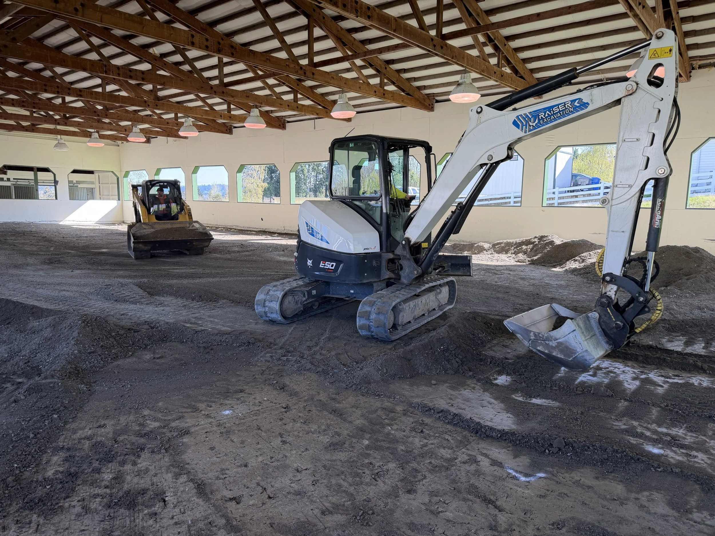 Construction site with a small excavator and compact loader inside a building with wooden roof trusses and large windows, with dirt and gravel on the ground.