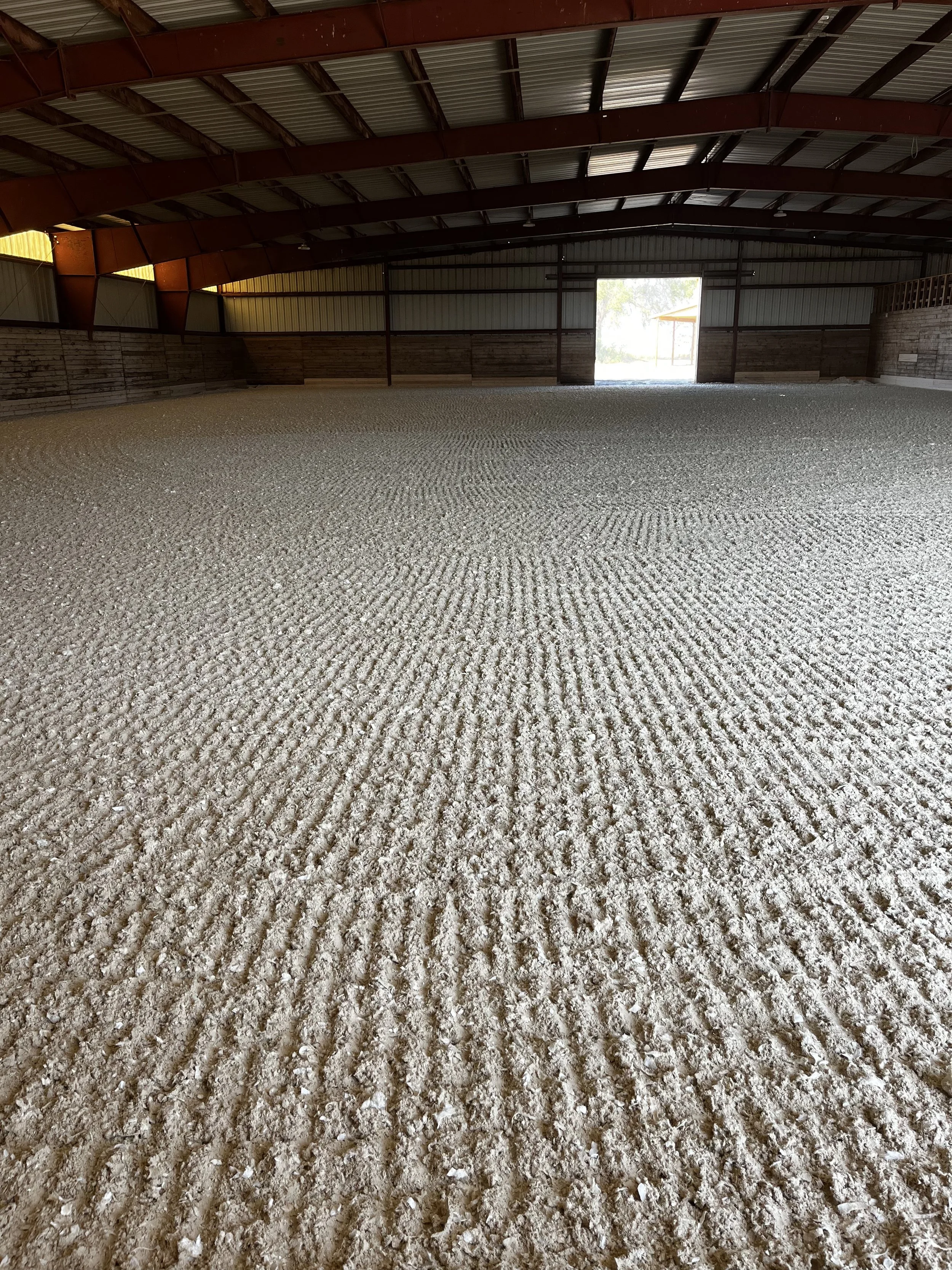 An indoor riding arena with a sand or dirt riding surface, wooden and metal siding and roof, and an opening at the far end allowing natural light to enter.
