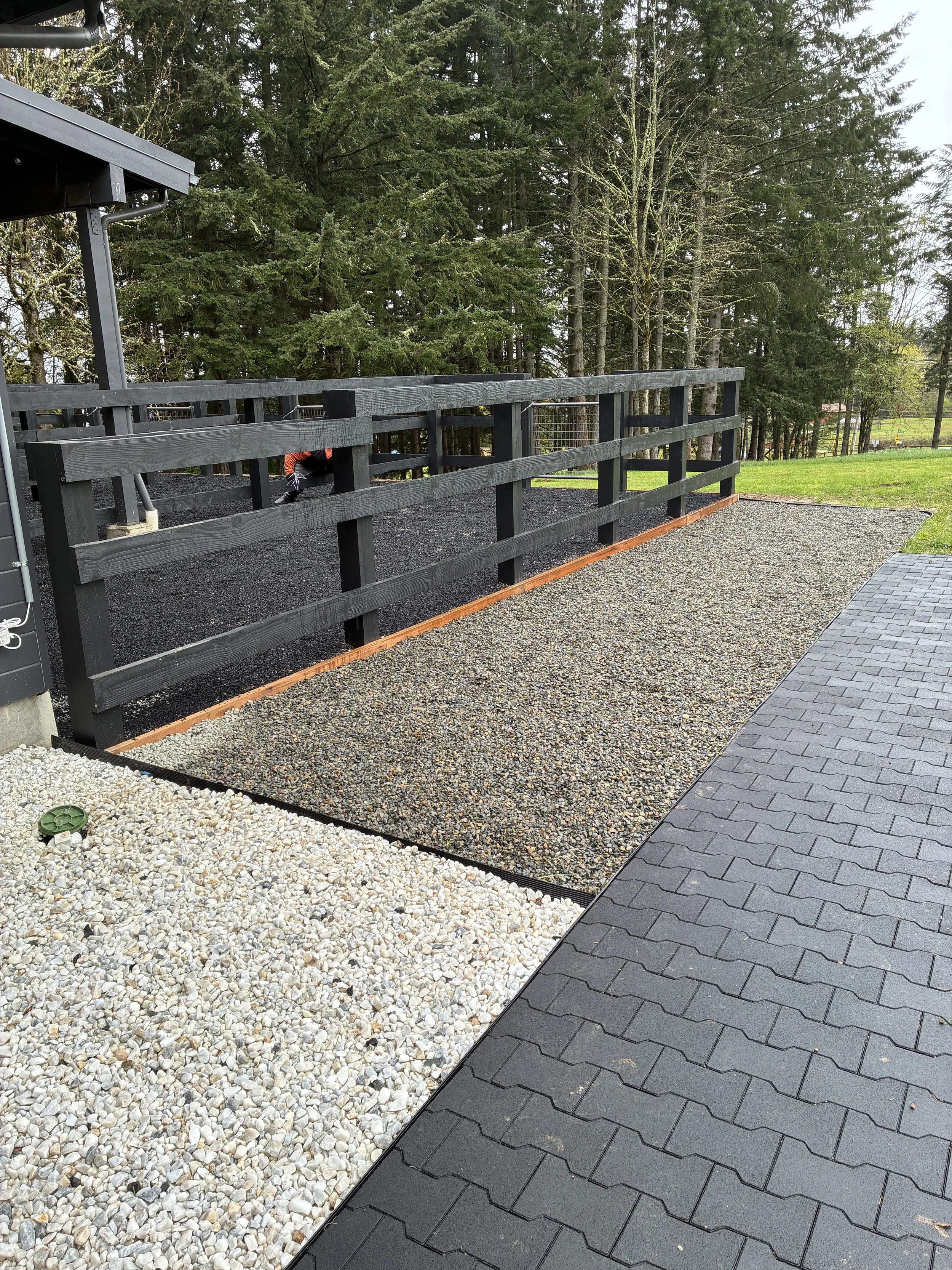 A backyard patio area with different ground surfaces, including gravel, pavers, and dirt, bordered by a black wooden fence and surrounded by trees.