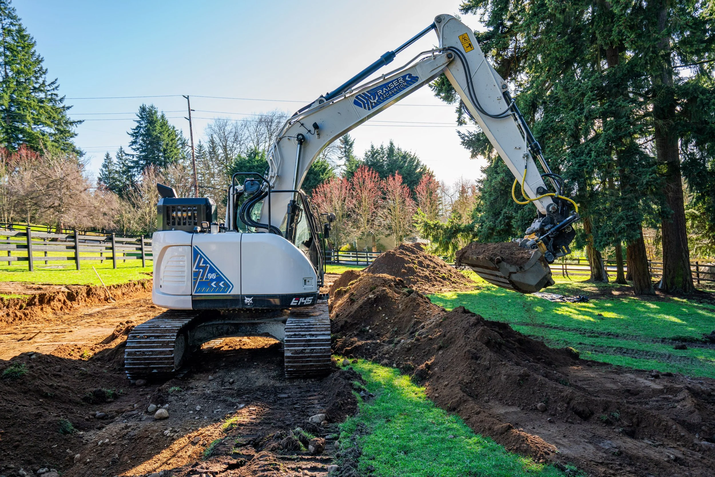 A white excavator with a black cab and tracks digging into the ground in a grassy yard surrounded by tall trees and a wooden fence.