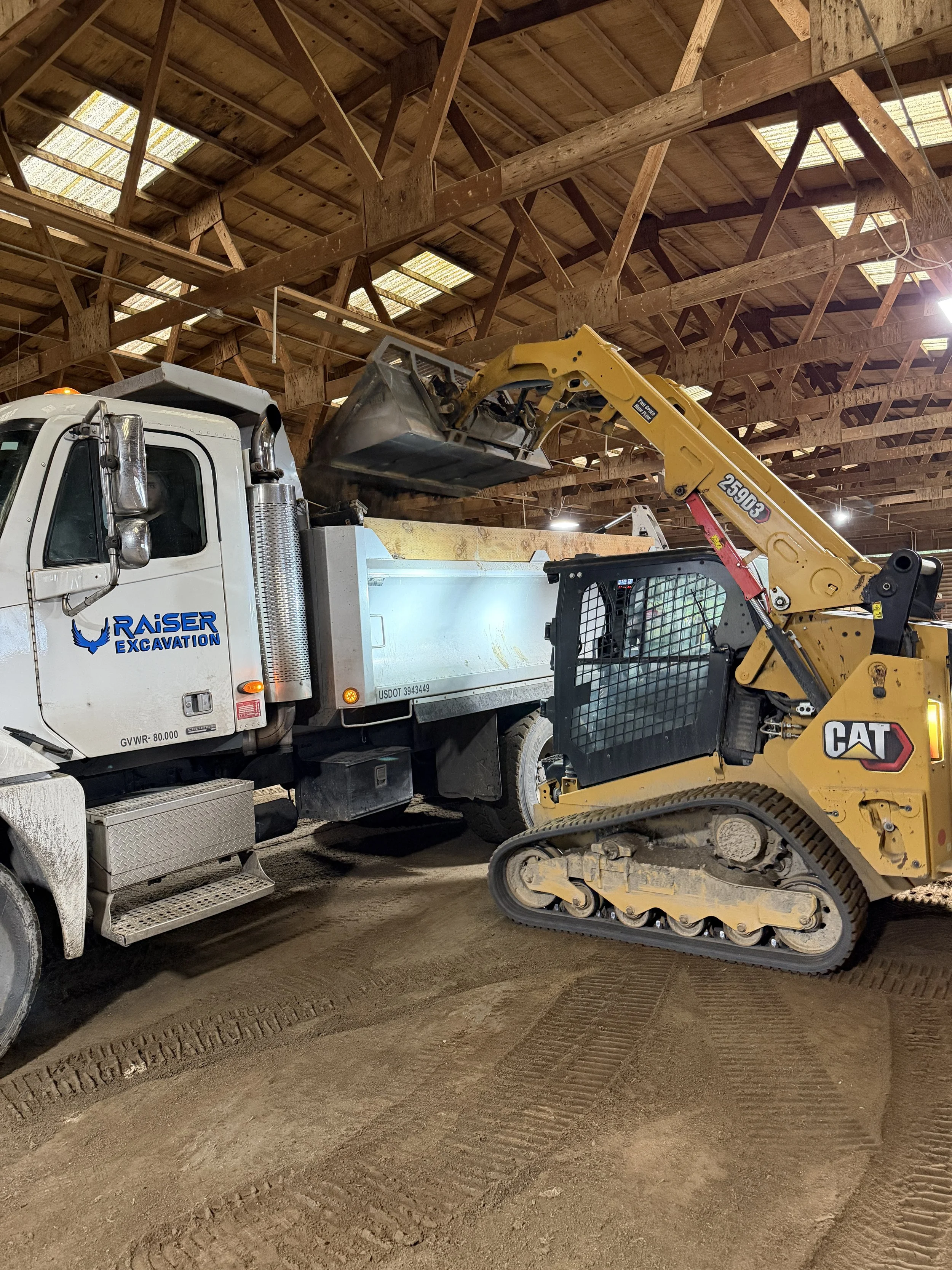 A construction site with a large white truck labeled 'RAISER EXCAVATION' and a yellow CAT tracked backhoe loader inside a wooden building under construction.