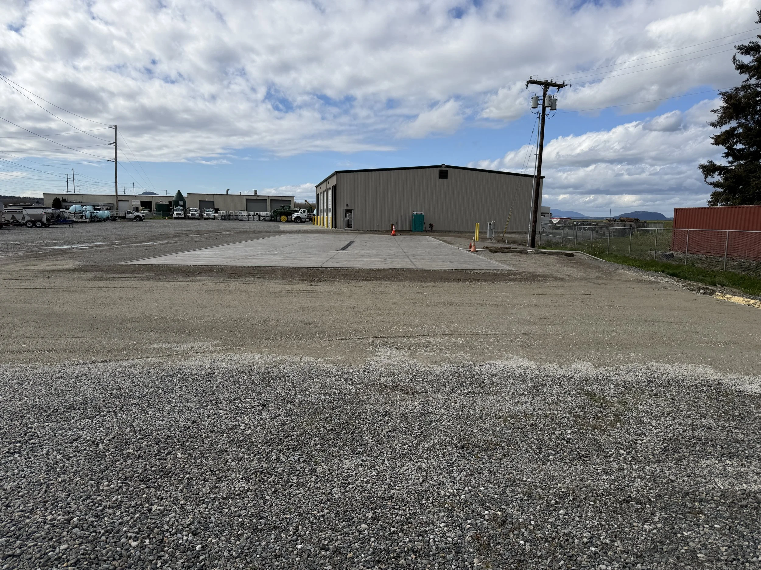 Industrial yard with large metal buildings, parked trucks, gravel ground, utility poles, fencing, and a partly cloudy sky.