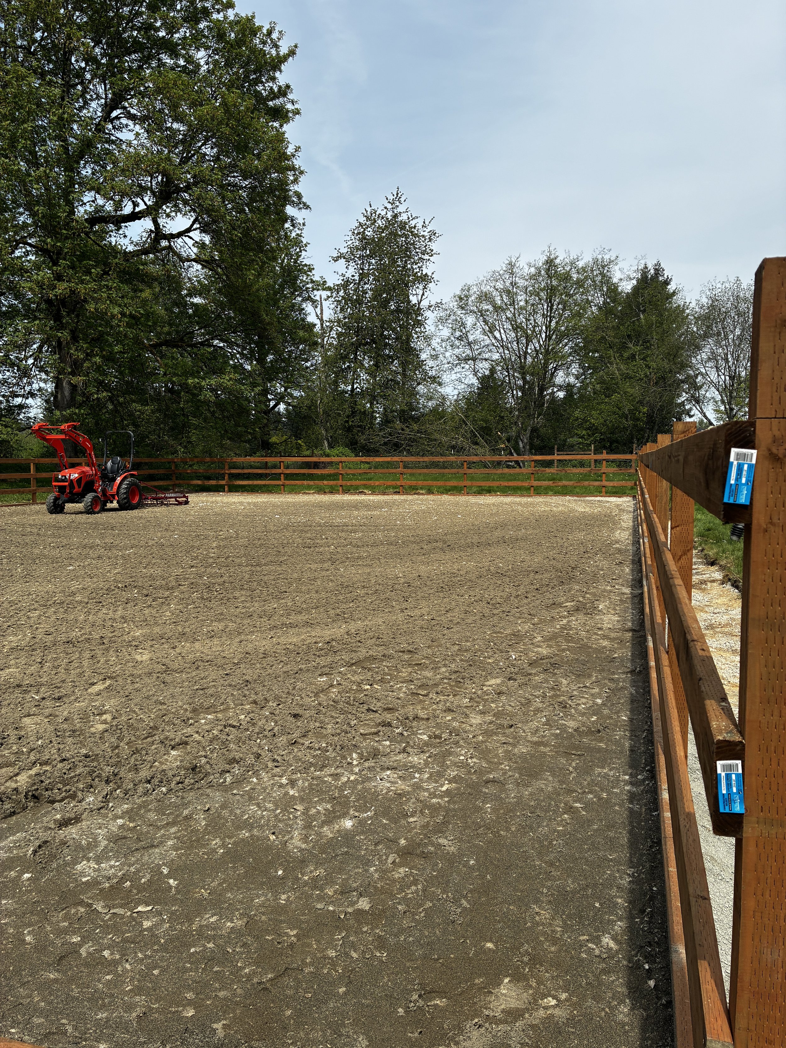 A red compact tractor with a front loader attachment and a mower or tiller in a fenced outdoor area, surrounded by trees under a cloudy sky.