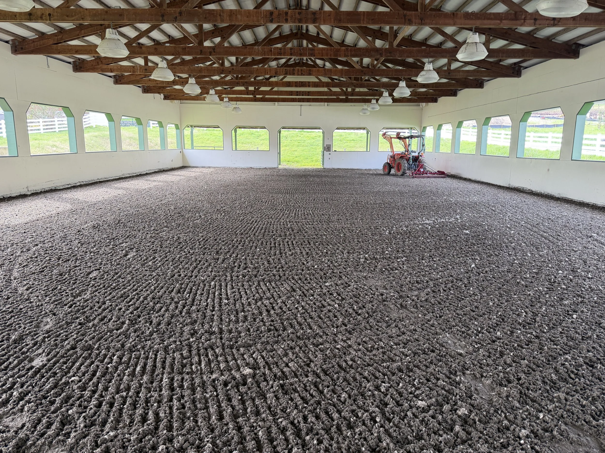 This is an indoor arena with a gravel floor, wooden roof beams, and windows around the perimeter. There is a small tractor with a red attachment inside the arena.