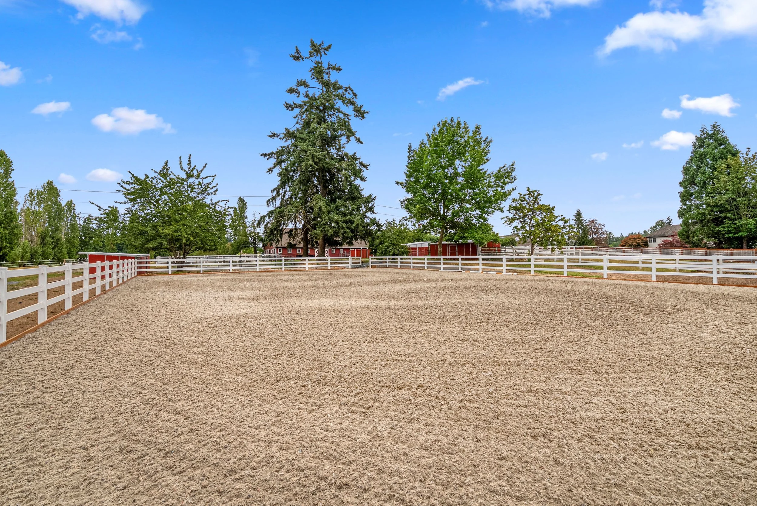 An outdoor riding arena with a dirt riding surface, white fencing, trees, and a red barn or stable in the background under a blue sky with some clouds.