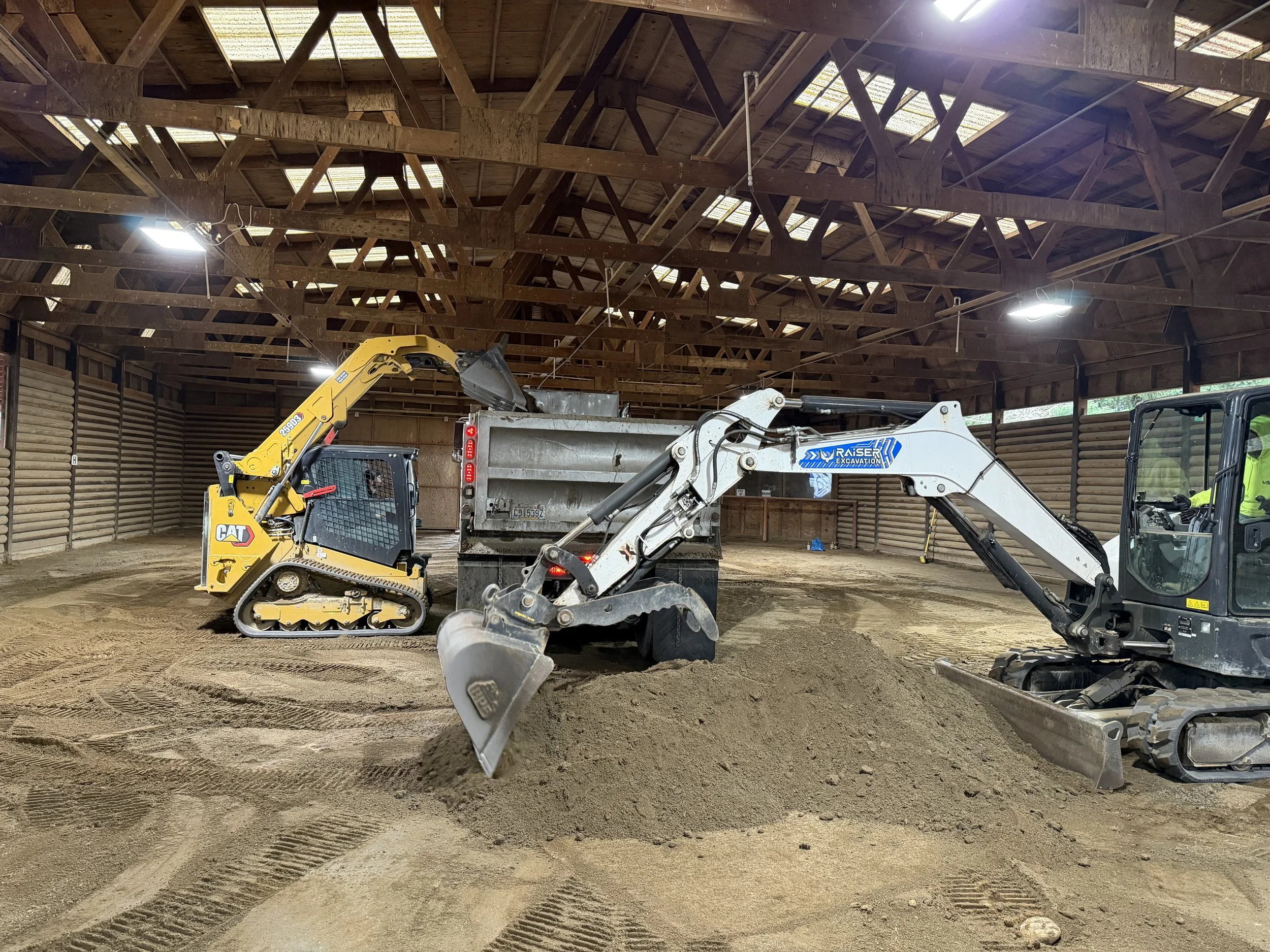 Interior view of a wooden barn under construction with construction equipment including a small excavator and a dump truck on a dirt floor.