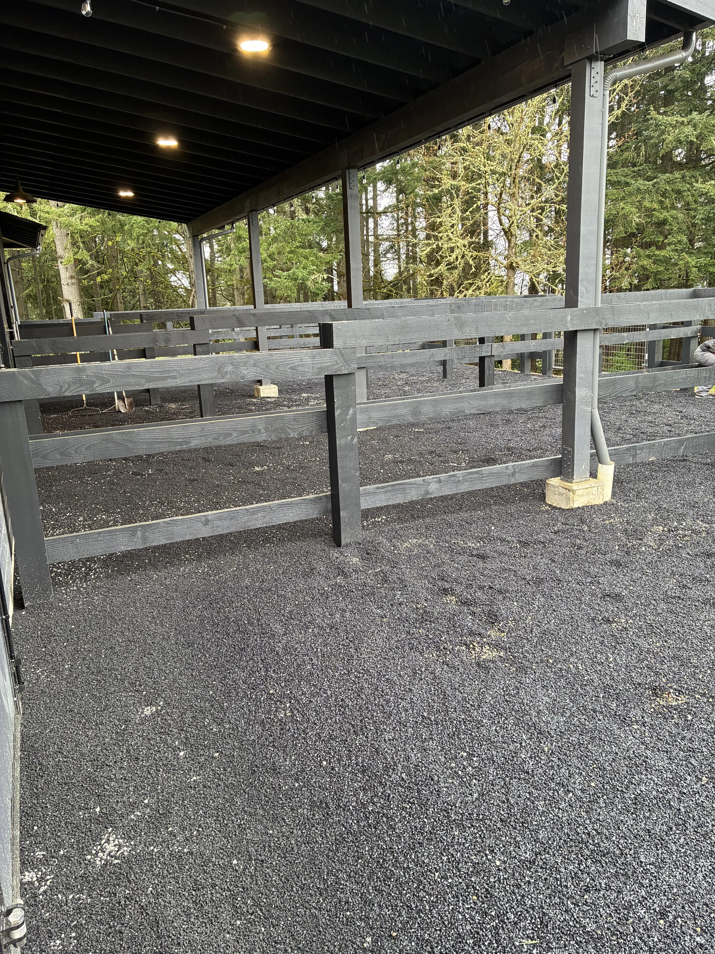 Outdoor partially covered area with black wooden railing and ground surface made of small black and white gravel. Trees are visible in the background.
